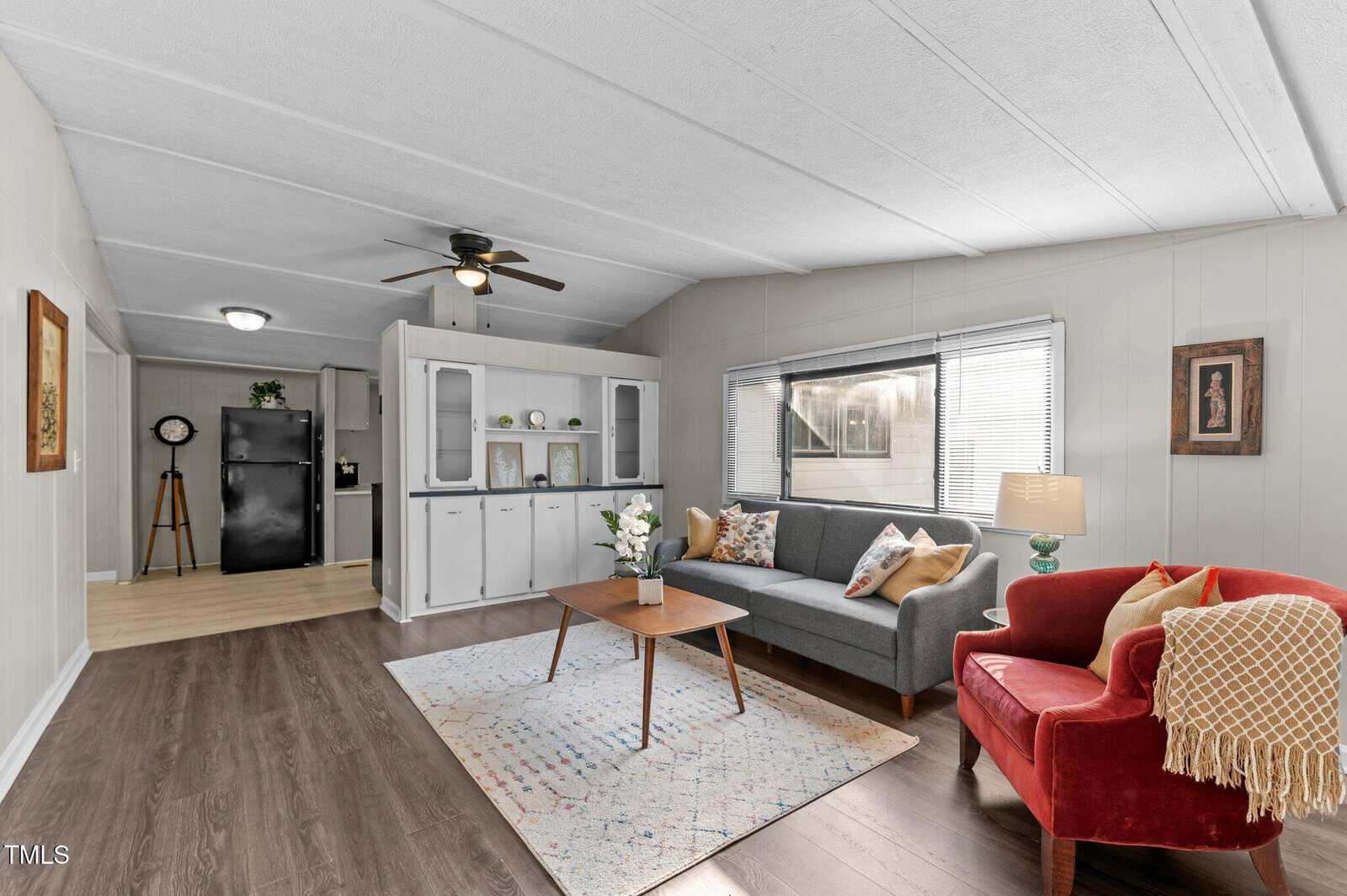 A bright living room with a gray sofa, red armchair, wooden coffee table, and patterned rug. Built-in white cabinets line one wall, and a kitchen with black appliances is visible in the background.
