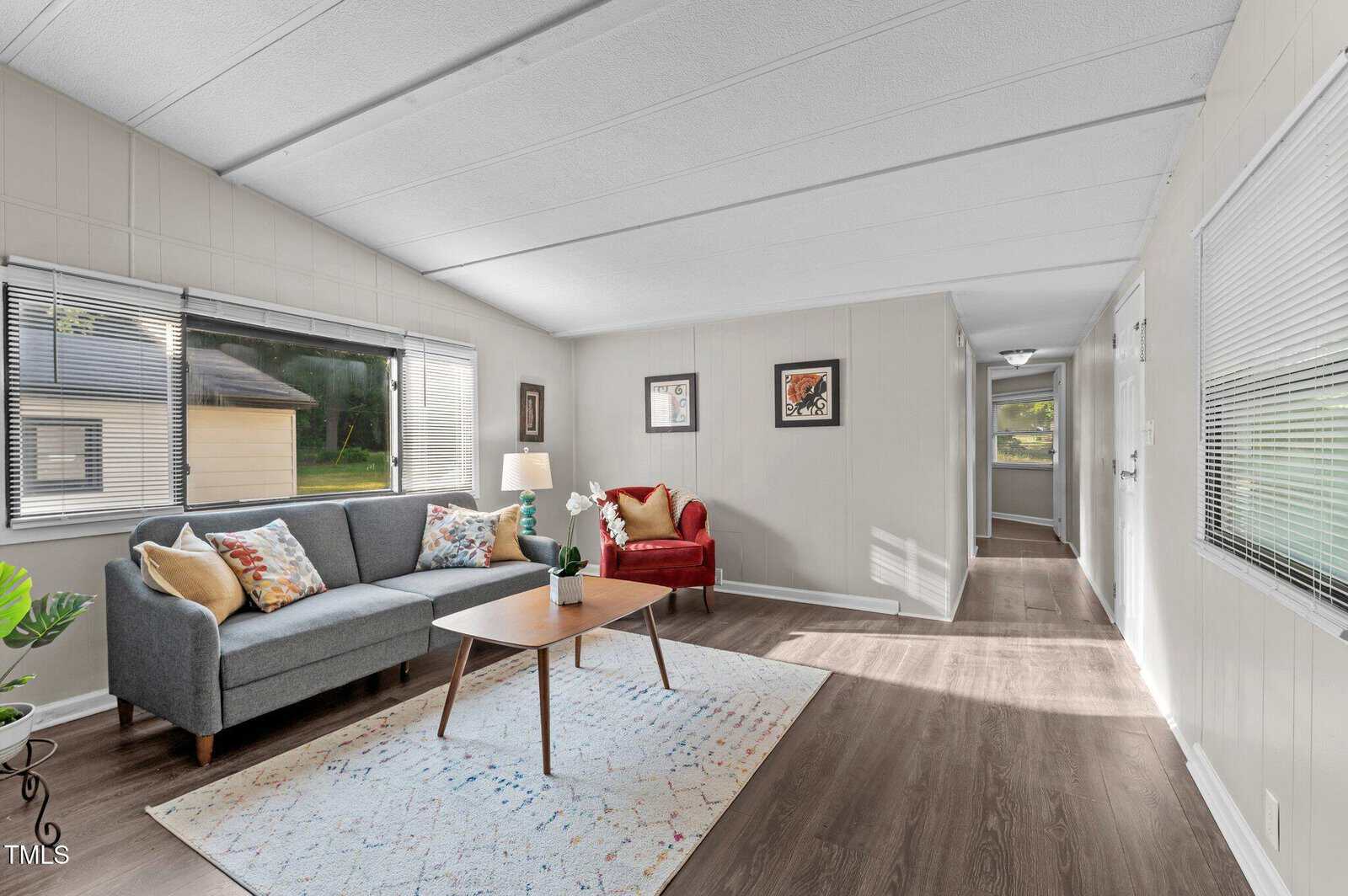 A simple living room with a gray sofa, red armchair, wooden coffee table, rug, and two windows. Walls are light-colored, floor is wood, and framed art hangs on the walls. Sunlight streams through the windows.