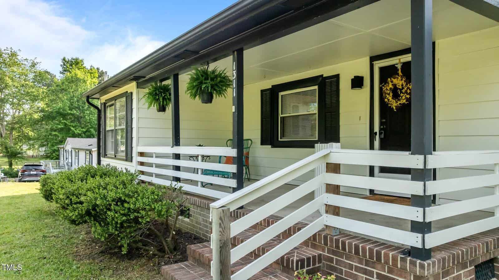 Single-story house with white siding and black trim, a covered front porch with white railings and brick steps, hanging ferns, a wreath on the door, and bushes lining the yard under a partly cloudy sky.