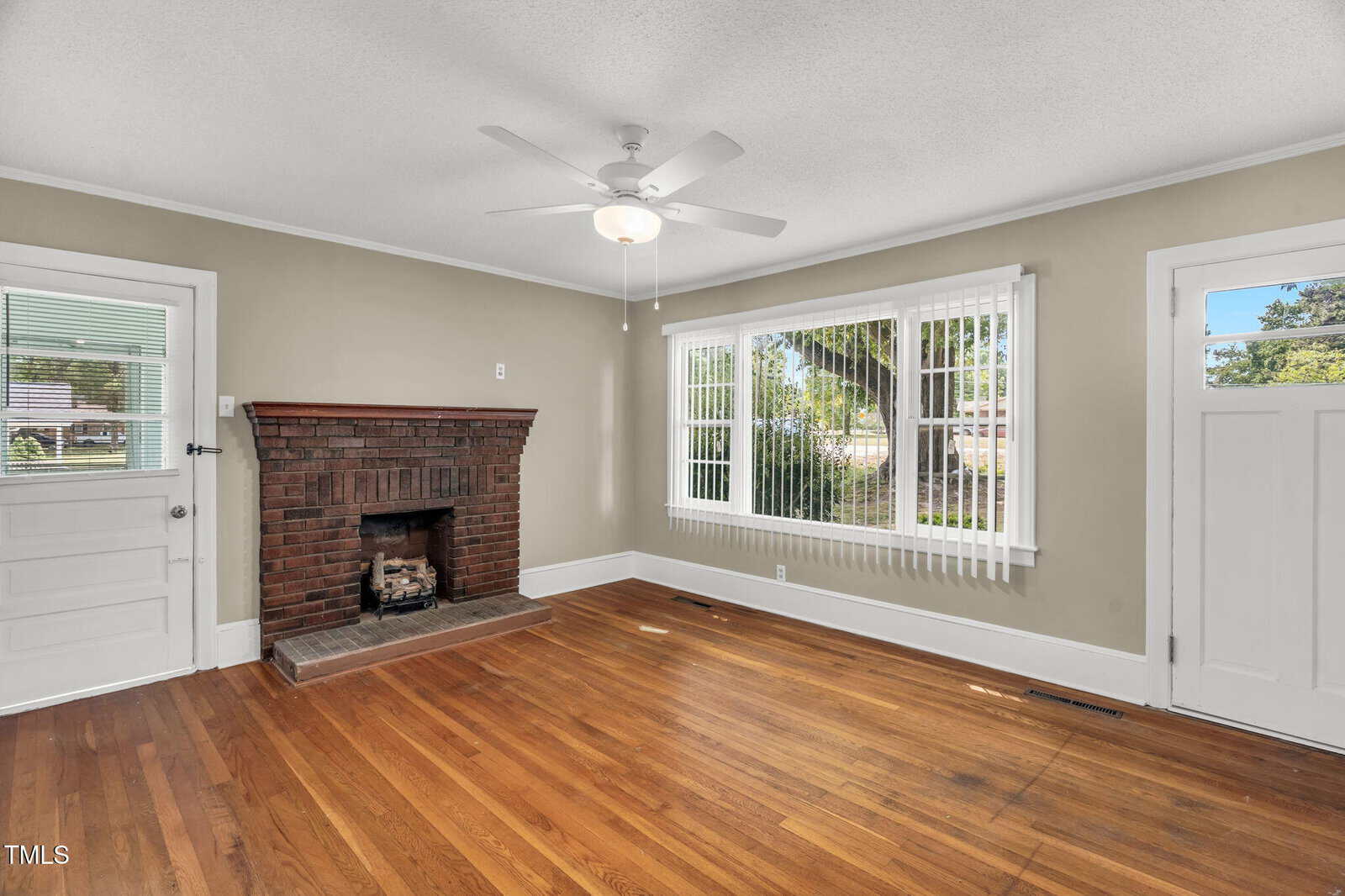 A living room with hardwood floors, a brick fireplace, beige walls, a ceiling fan, vertical blinds on a large window, and a white door leading outside. Natural light fills the space.