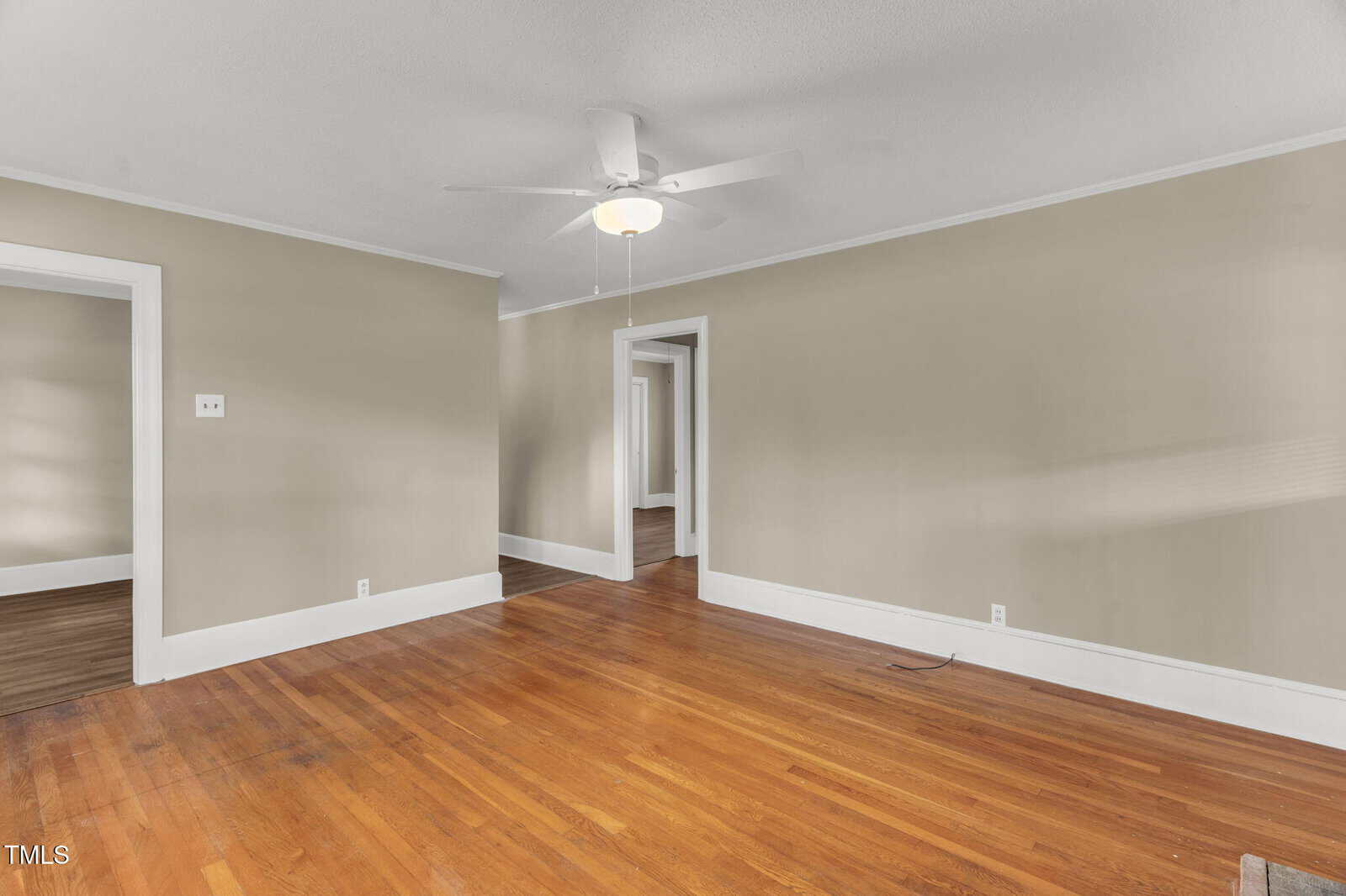 A room with beige walls, white trim, and a wooden floor. There is a ceiling fan with a light, and three doorways leading to other rooms. The space is empty and well-lit by natural light.