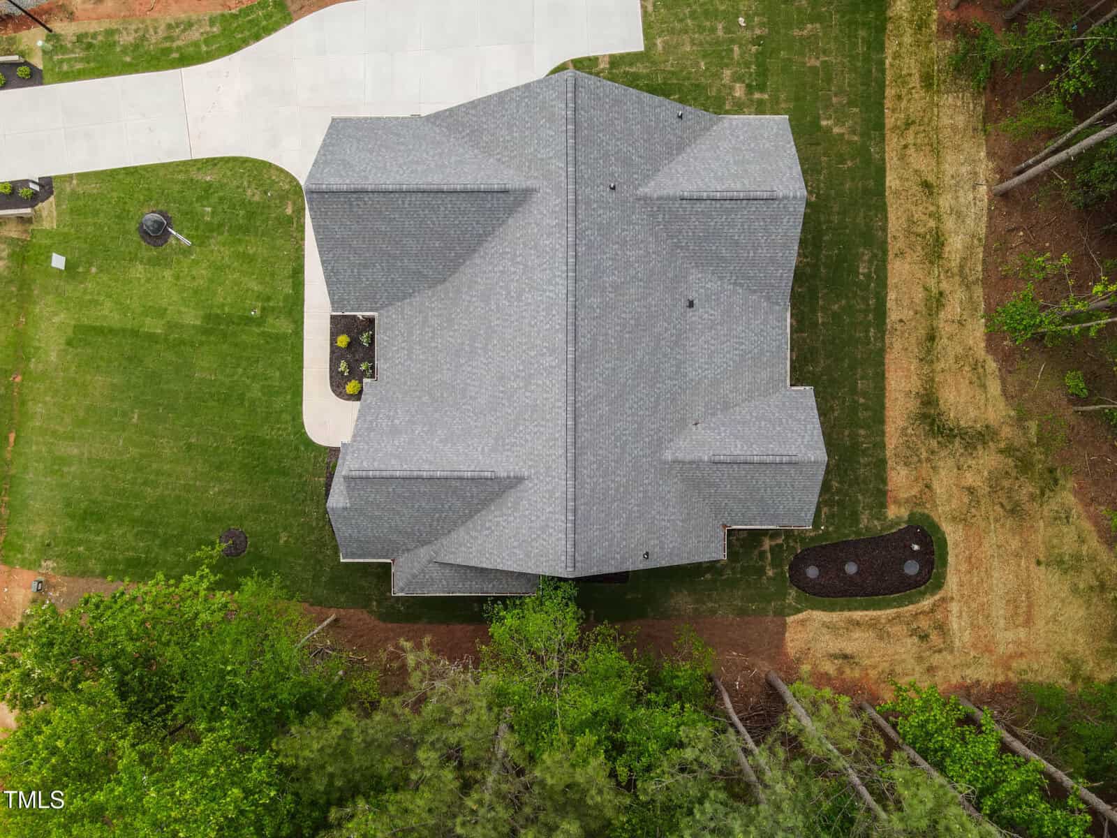 Aerial view of a house with a gray roof, surrounded by green grass, a driveway on the left, landscaped areas with mulch, and trees bordering the bottom of the image.