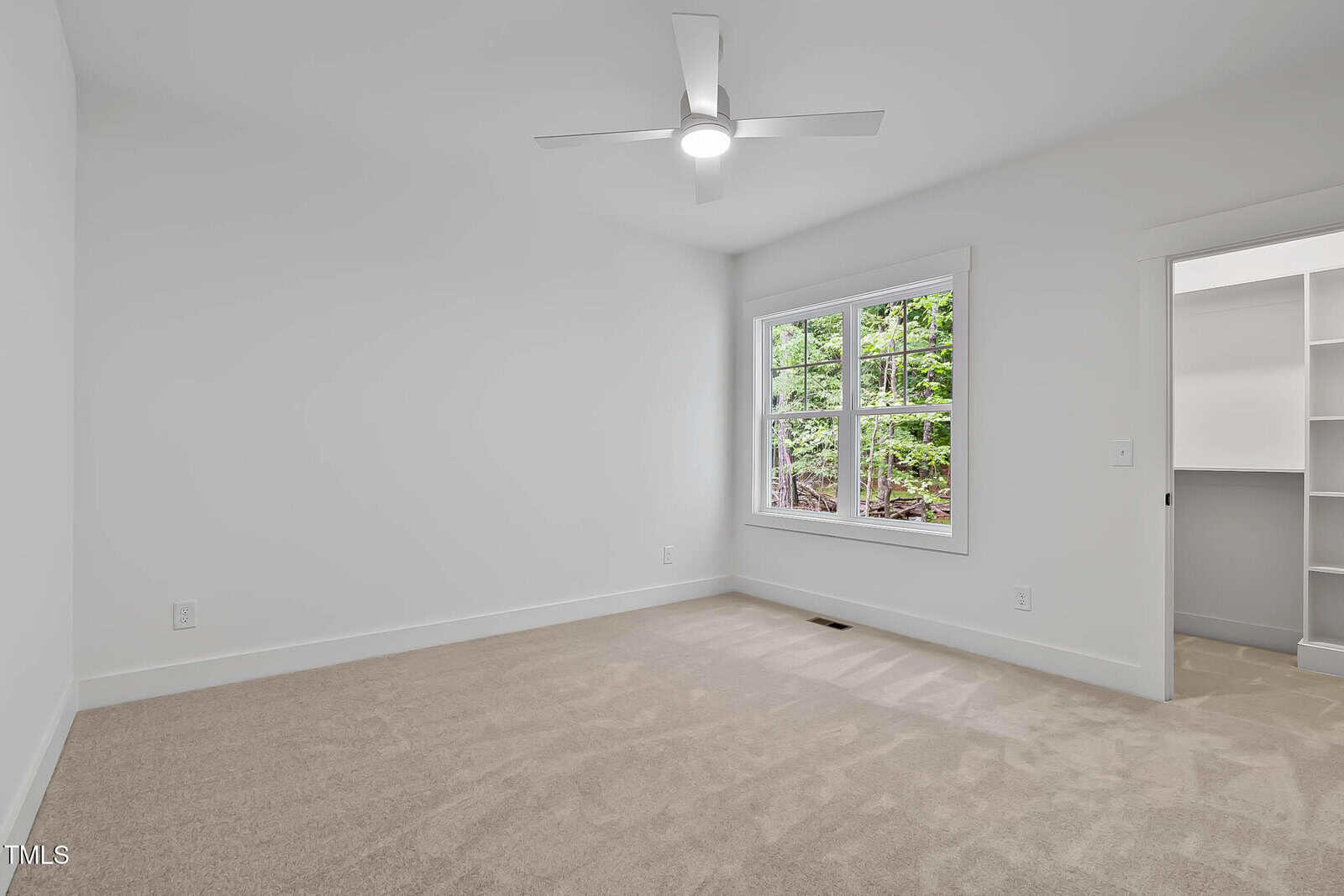 A bright, empty room with beige carpet, white walls, a ceiling fan with a light, a large window showing greenery outside, and an open doorway leading to a walk-in closet with shelves.