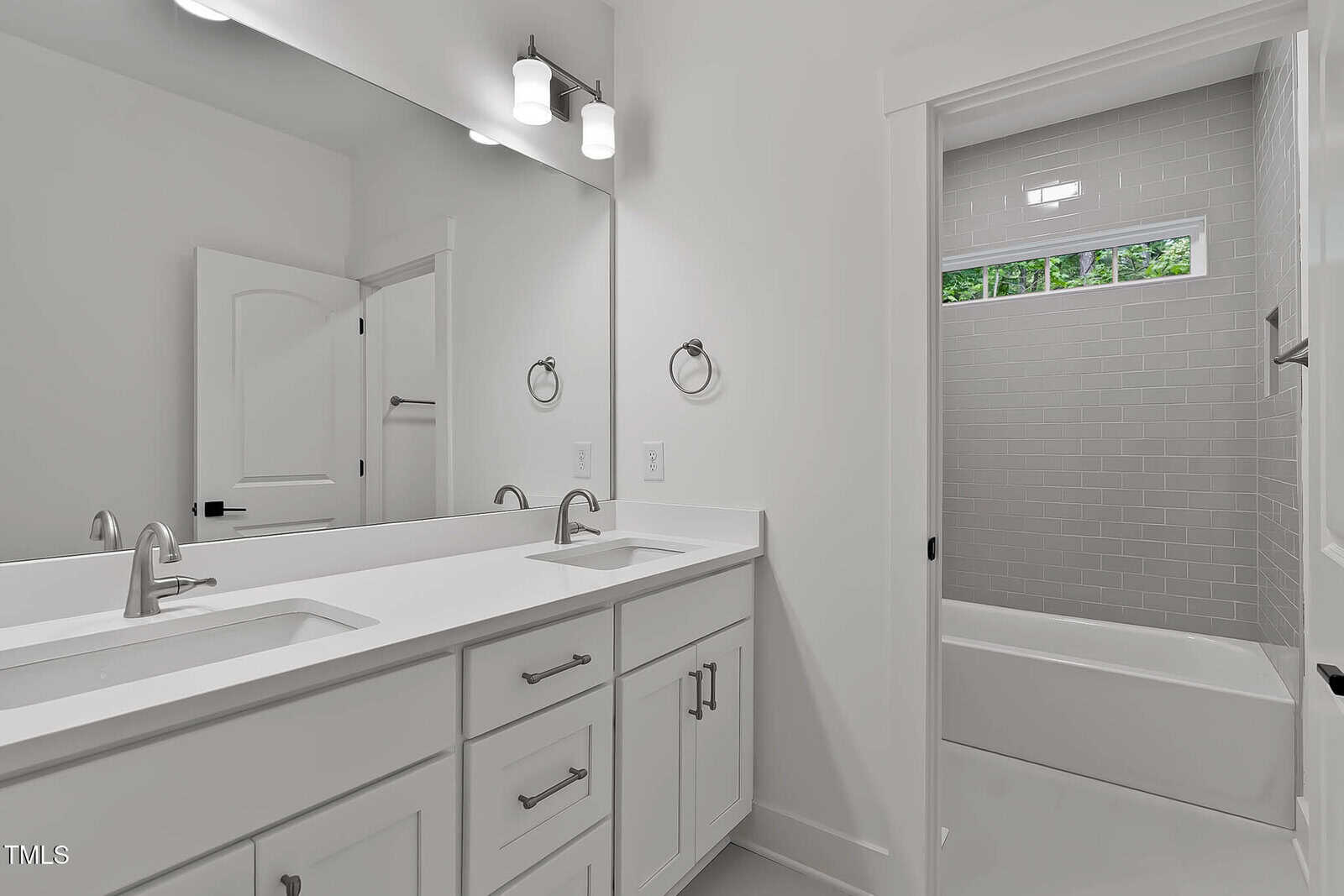 A modern bathroom with a double sink vanity, white cabinets, two mirrors, silver fixtures, and a bathtub with light gray tile surround. The room features white walls and a door leading to the bathtub area.