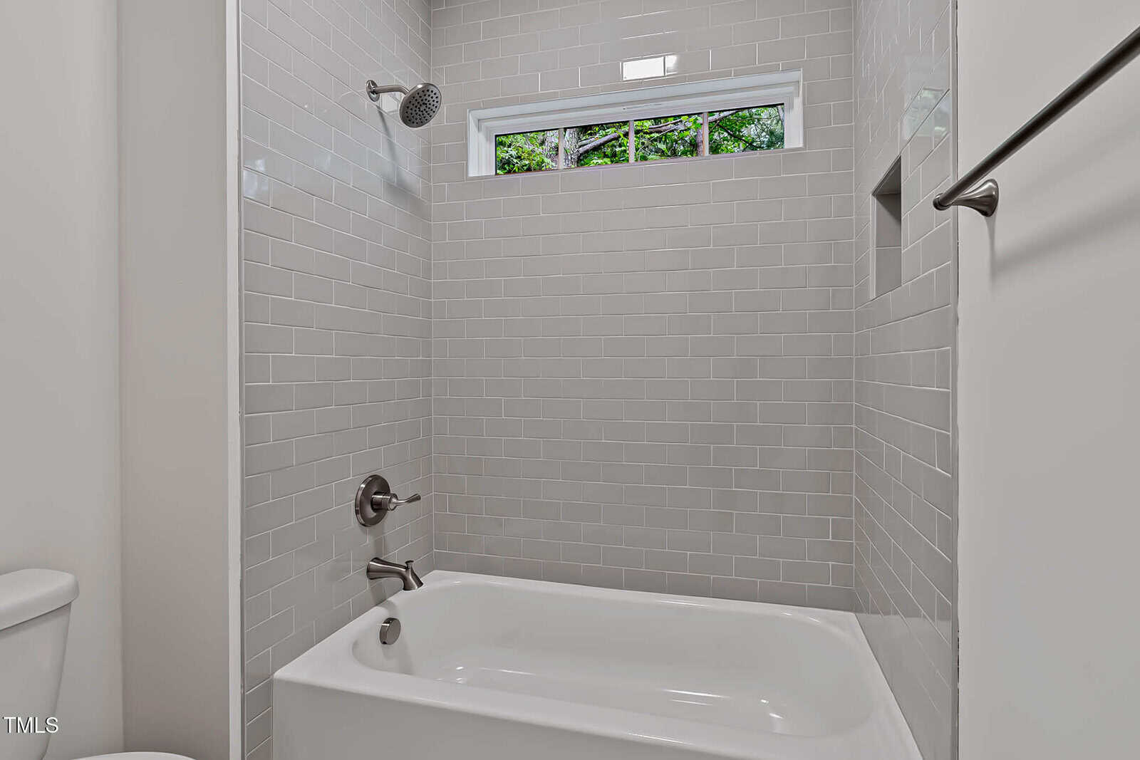 A white bathtub with silver fixtures is set against a wall of glossy light gray subway tiles. There is a small horizontal window above the shower letting in natural light and a towel bar is mounted on the right wall.