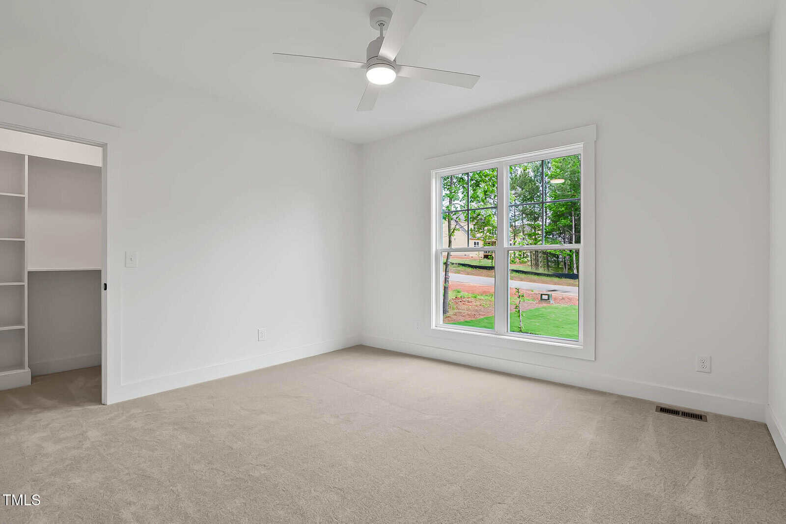 A bright, empty room with beige carpet, white walls, a ceiling fan, a large window overlooking a lawn and trees, and an open closet with built-in shelving.