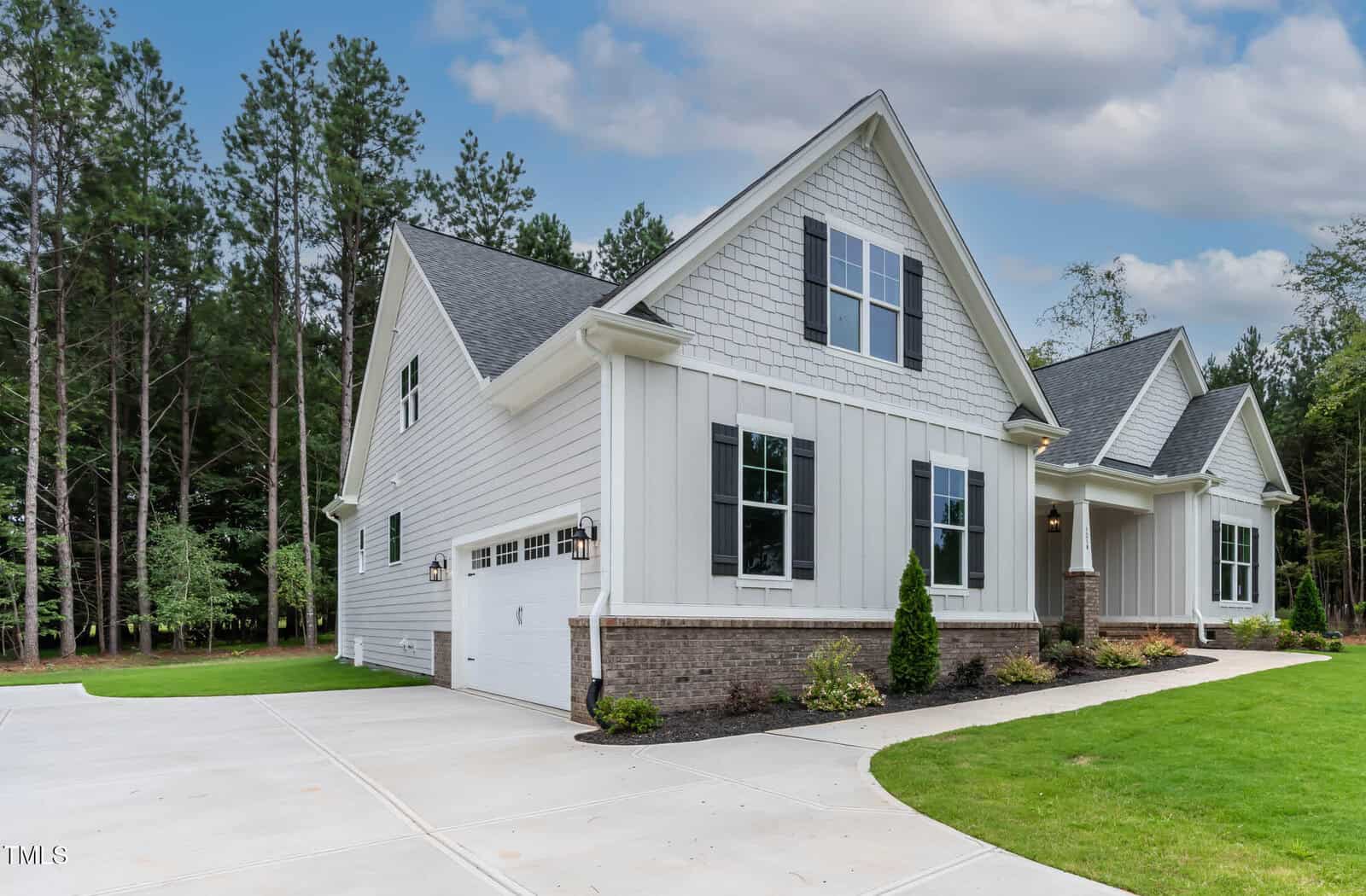A modern white house with black-trim windows, a gray shingle roof, and attached double garage sits next to a driveway and green lawn. Trees and a cloudy sky are visible in the background.
