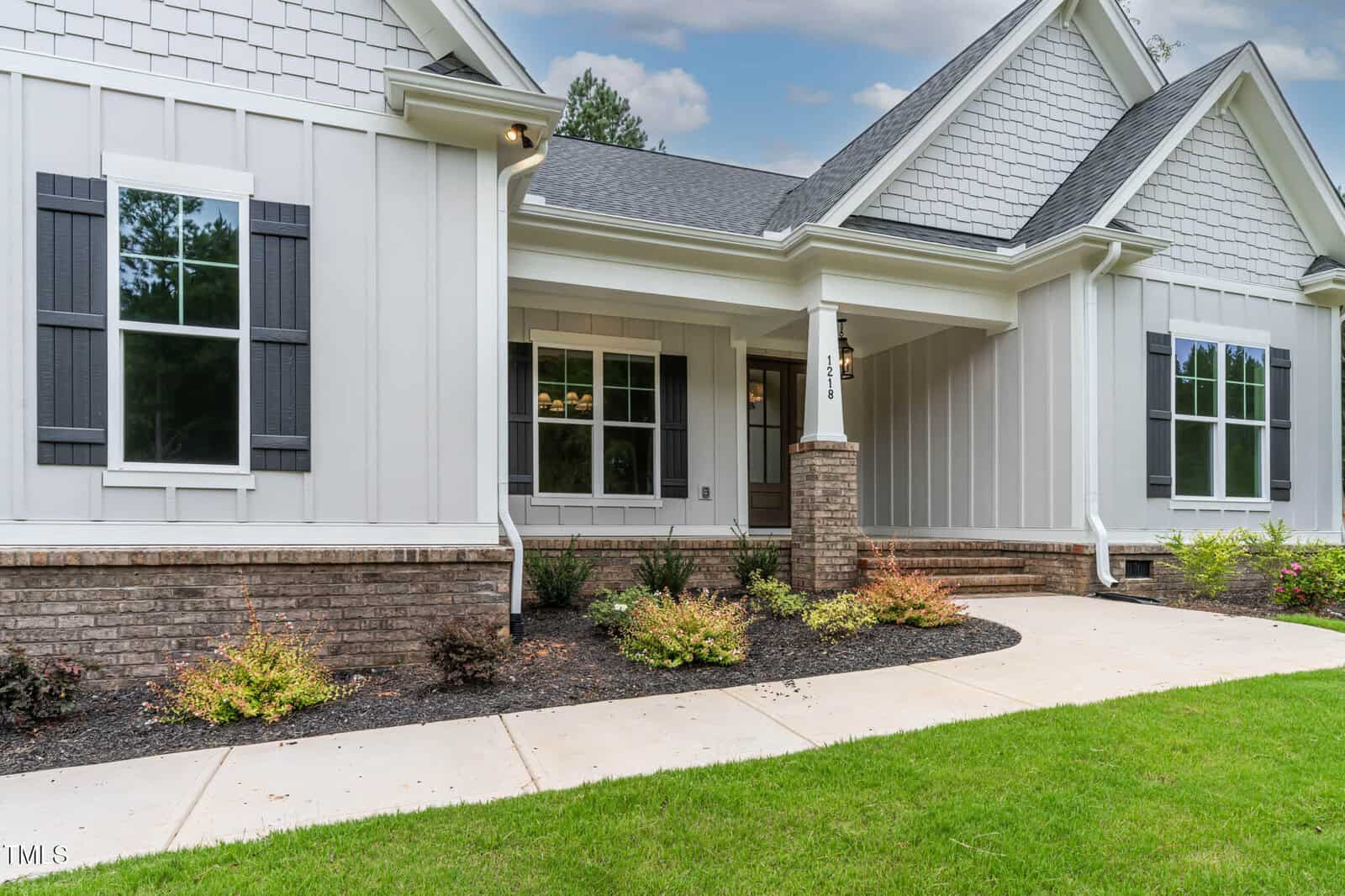 A modern single-story house with light gray siding, black shutters, and a brick foundation. There is a covered front porch with columns, a concrete walkway, and landscaped shrubs bordering the lawn.