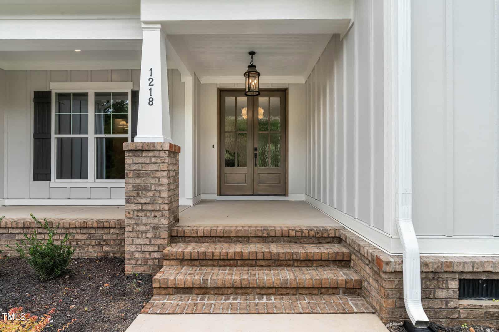 A front porch with three brick steps leading to double wooden doors, a hanging lantern light above, white vertical siding, a brick column, house number 1218, and a white downspout on the right.