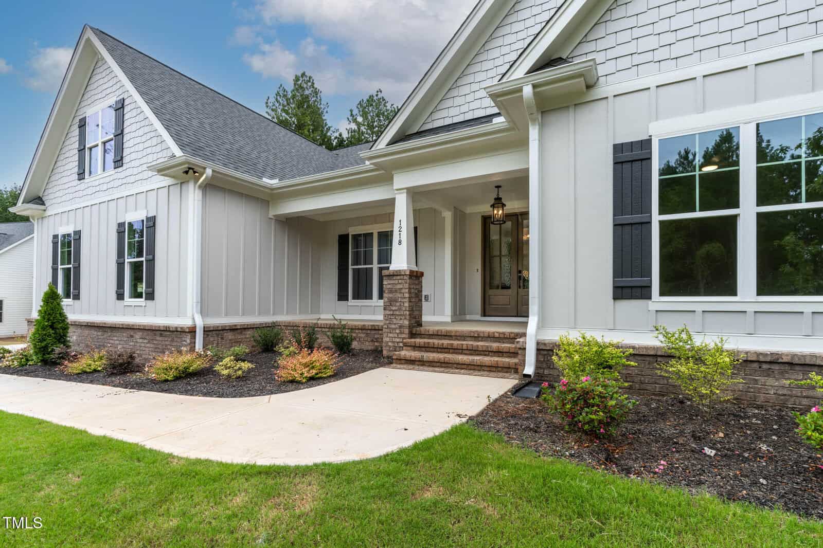 A modern single-story house with gray siding, white trim, black shutters, brick accents, and a covered front porch. Manicured bushes and a curved sidewalk lead to the entrance.