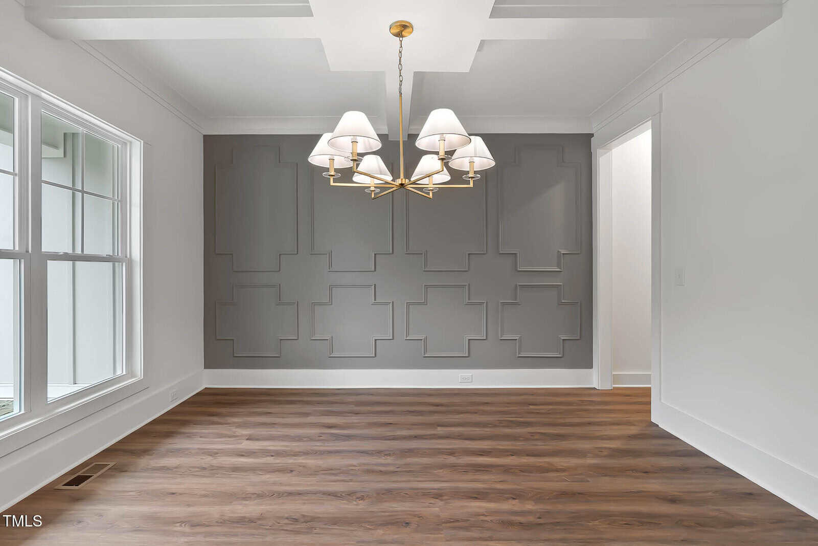 A dining room with a geometric accent wall painted gray, a gold chandelier with white lampshades, large windows on the left, and wood flooring. The room is empty with white walls and trim.