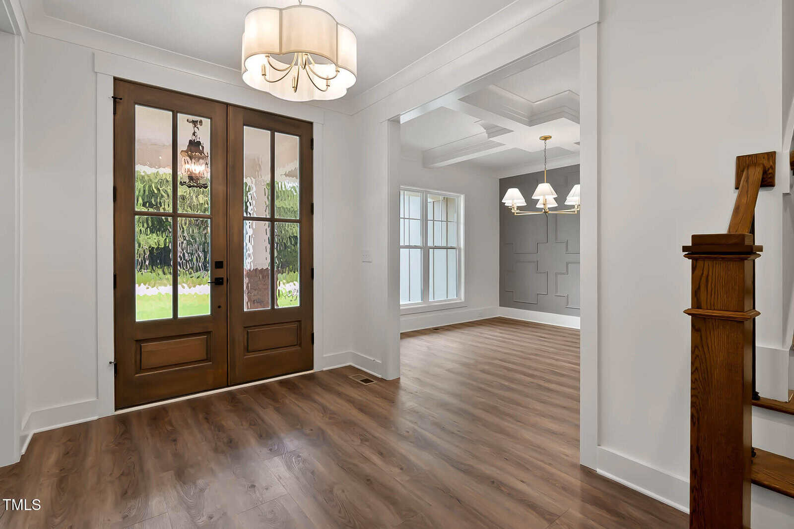 A bright entryway with double wooden doors, wood flooring, white walls, a modern chandelier, and a view into a formal dining room with large windows and another chandelier.