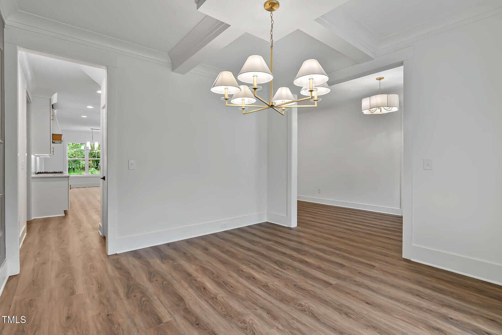A bright, empty room with white walls, a tray ceiling, wood-look floors, two modern chandeliers, and a view into a hallway leading to another well-lit living space with large windows.
