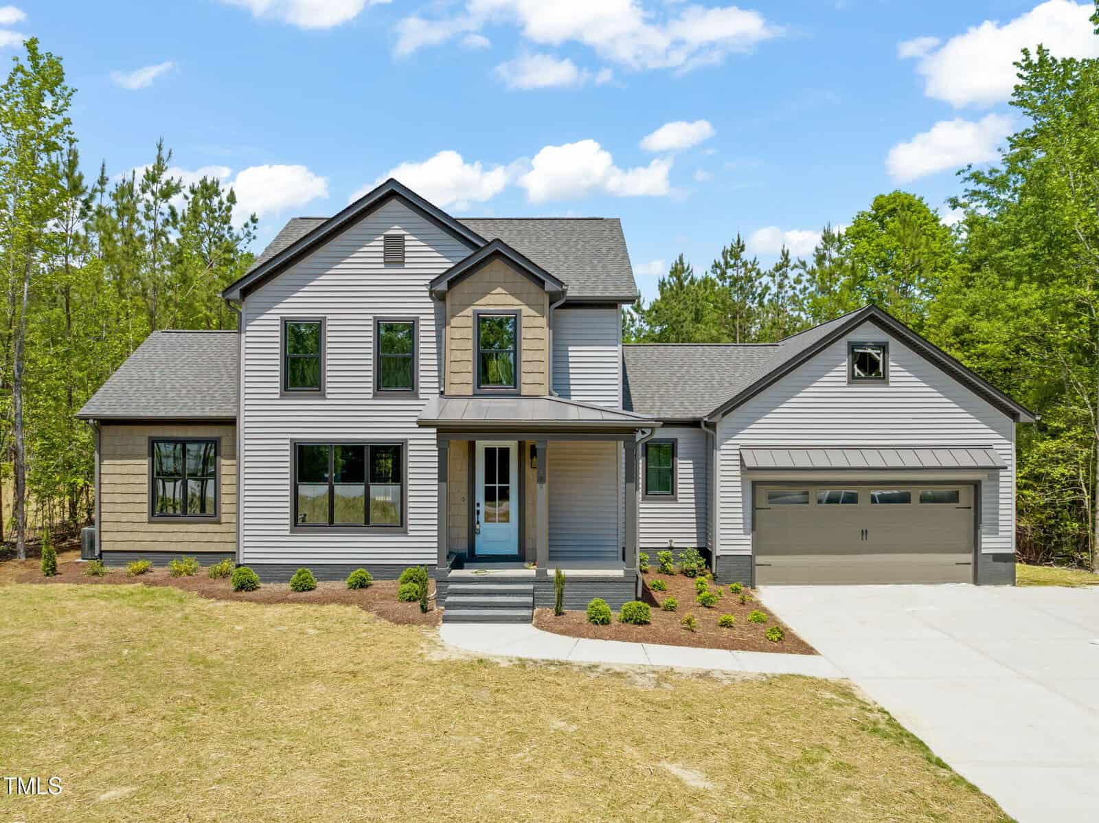 Two-story modern house with gray siding, multiple windows, a covered front porch, and attached two-car garage. The home is surrounded by trees and has a freshly landscaped yard with a concrete driveway.
