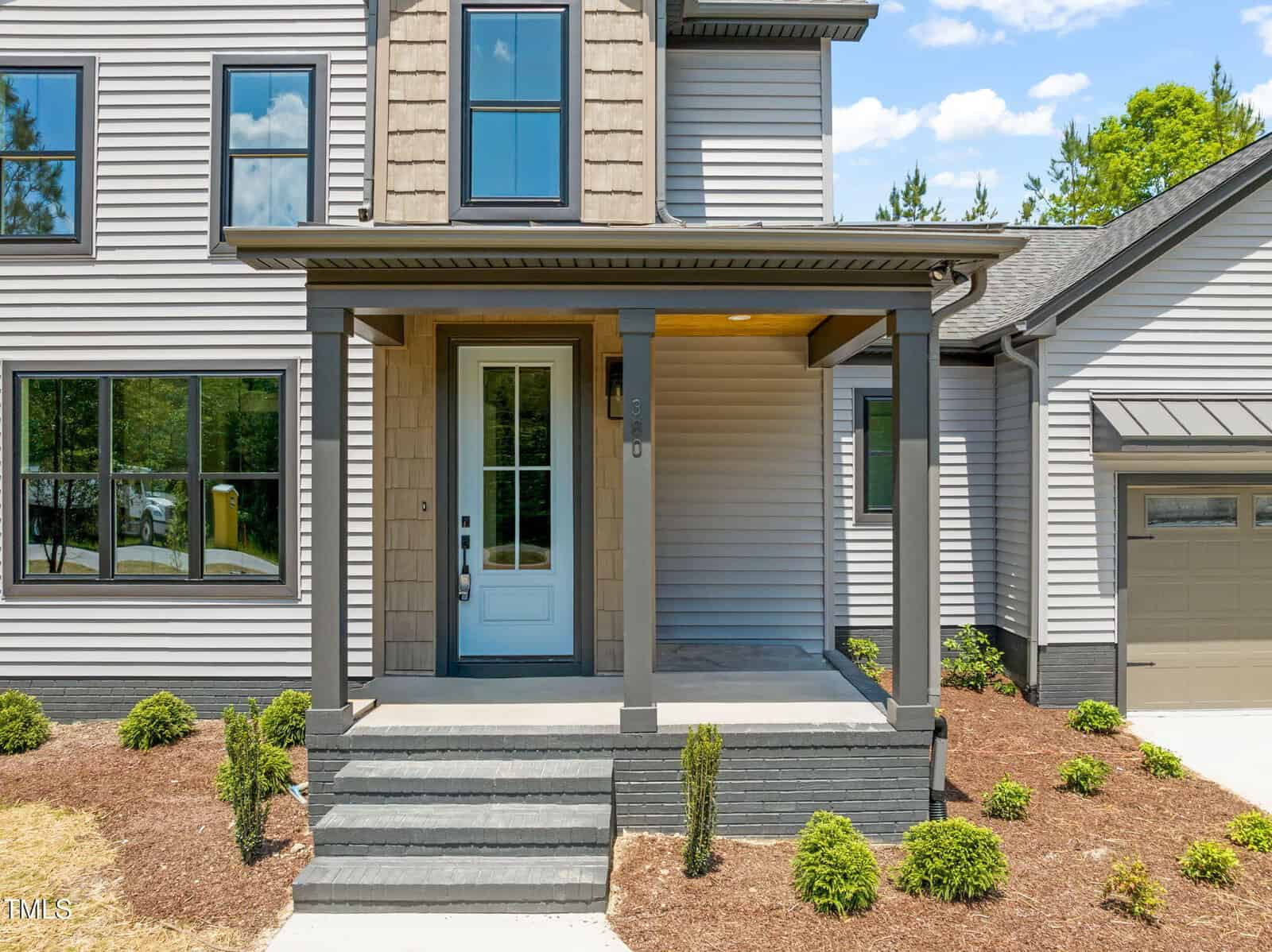 A modern house with gray siding and trim, featuring a small covered front porch with steps, a white front door, large windows, mulched landscaping, and a garage partially visible on the right.