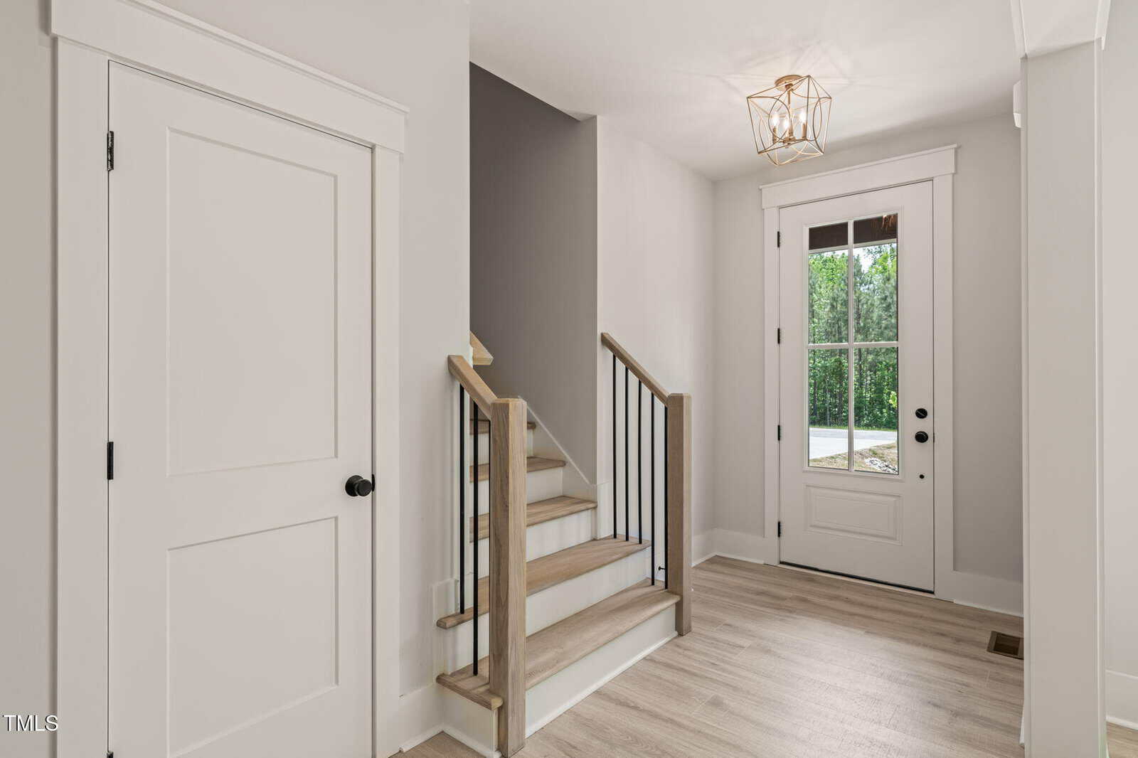 A modern entryway with light wood flooring, a white front door with glass panels, a small staircase with wood and metal railings, and neutral gray and white walls. A geometric light fixture hangs from the ceiling.