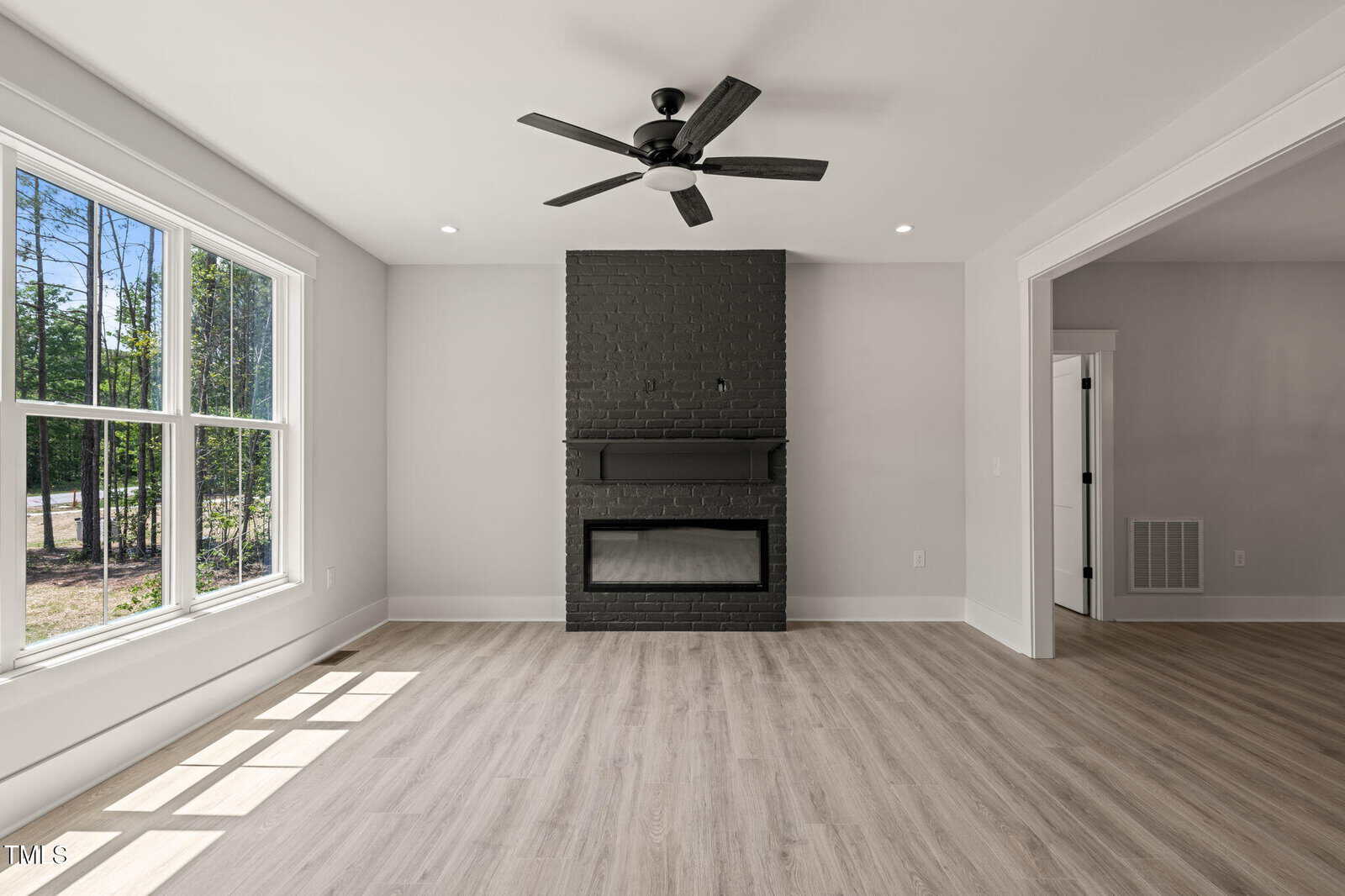 A bright living room with light wood flooring, large windows, and a black painted brick fireplace with a black ceiling fan above. White walls and trim complete the modern, minimal decor.