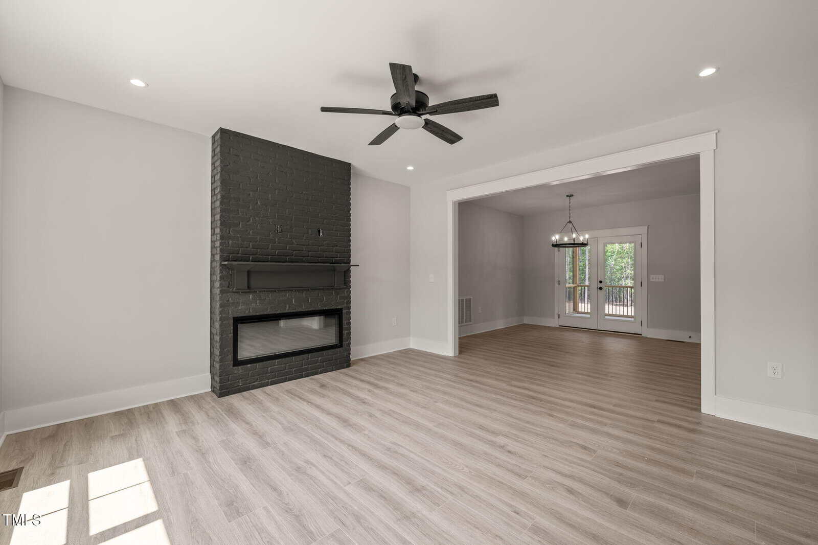 A modern living room with light wood flooring, a black ceiling fan, and a black painted fireplace. The room opens to a dining area with a large window and a glass door leading outside. The walls are painted light gray.