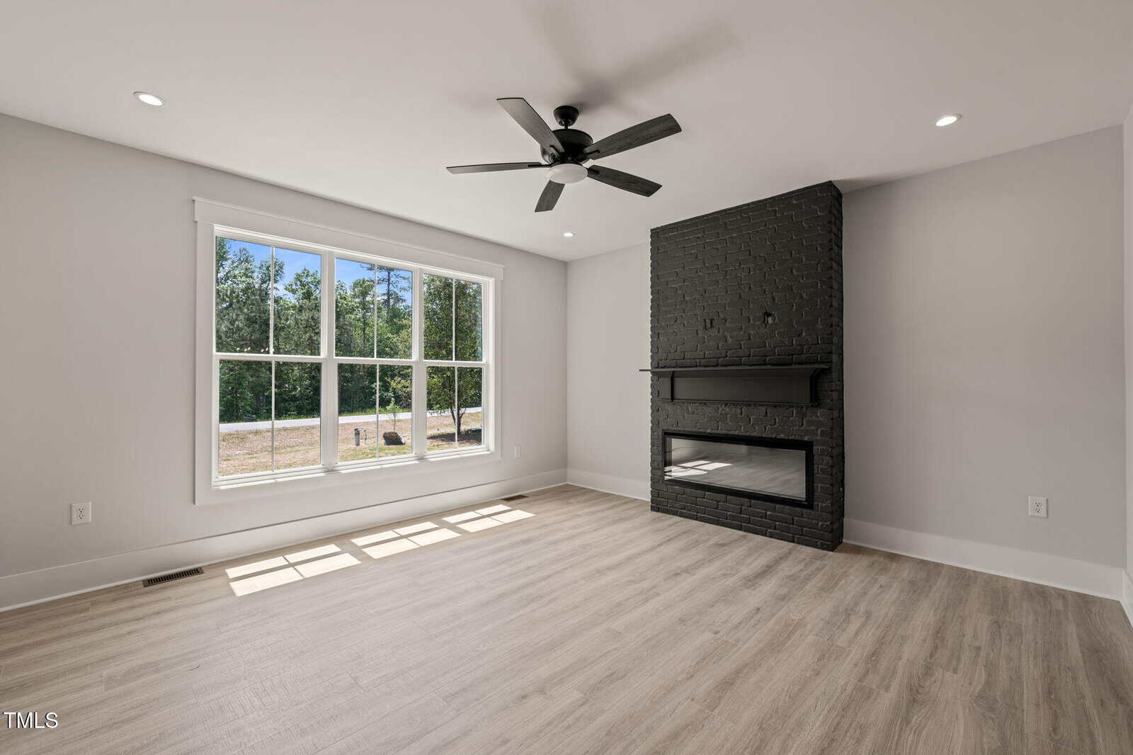 Bright, unfurnished living room with large window, light wood flooring, a ceiling fan, and a black brick fireplace with mantel against a white wall. Outdoors with trees visible through the window.