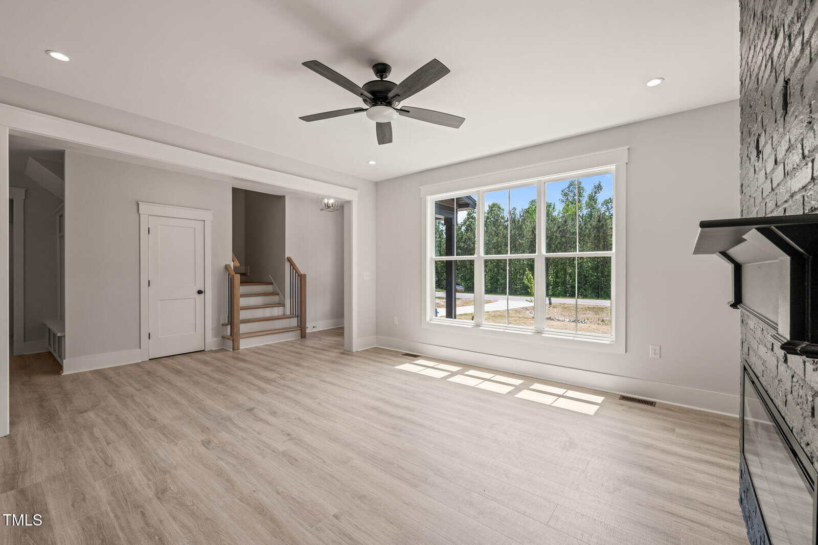 Bright living room with light wood flooring, a large window, white walls, a ceiling fan, and a partial view of stairs and fireplace. Sunlight streams in, illuminating the open space.