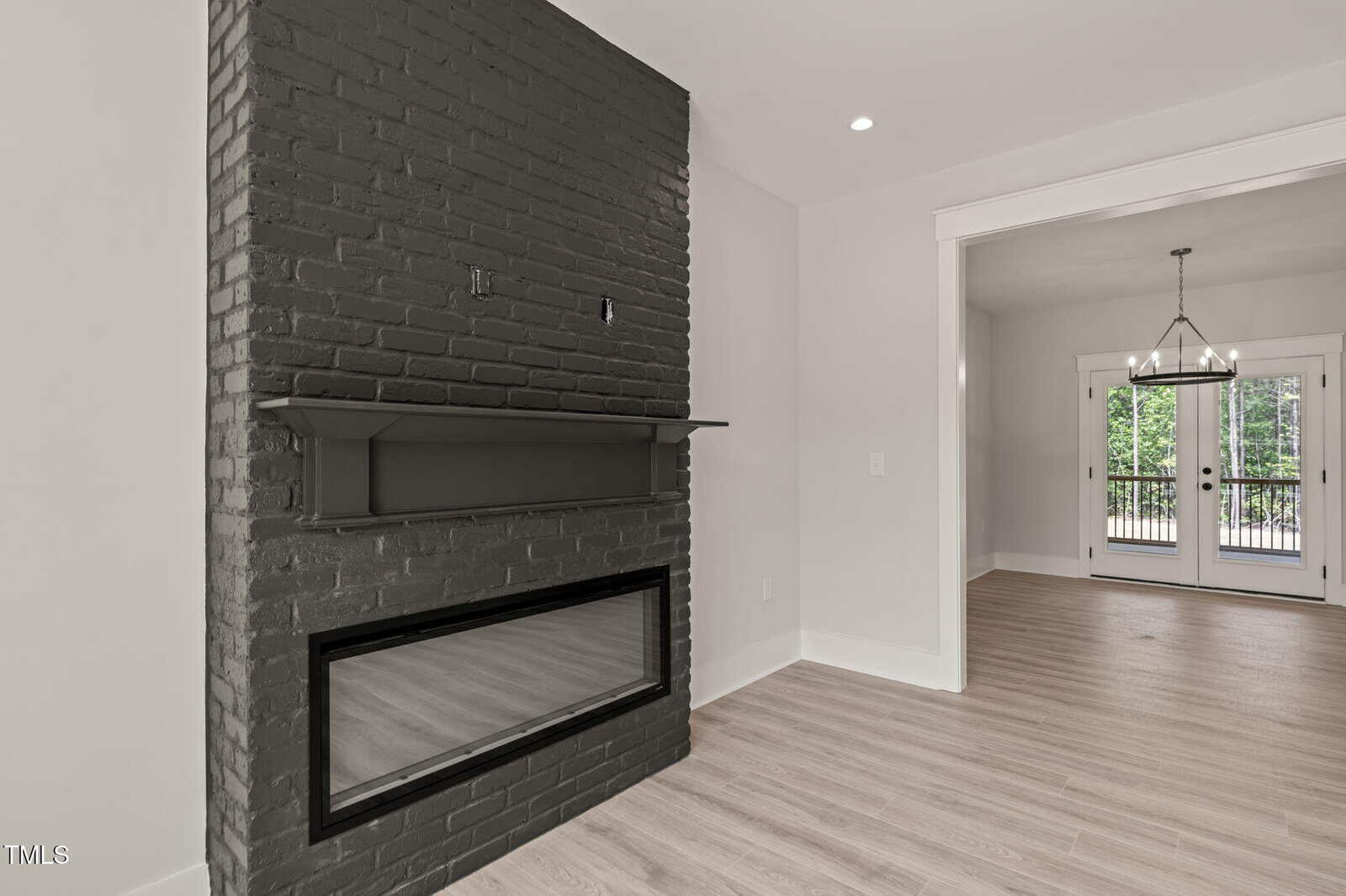 A room with light wood flooring, white walls, and a black brick fireplace. A shelf is above the fireplace. Double glass doors and a light fixture are visible in the adjacent dining area. Trees are seen through the doors.