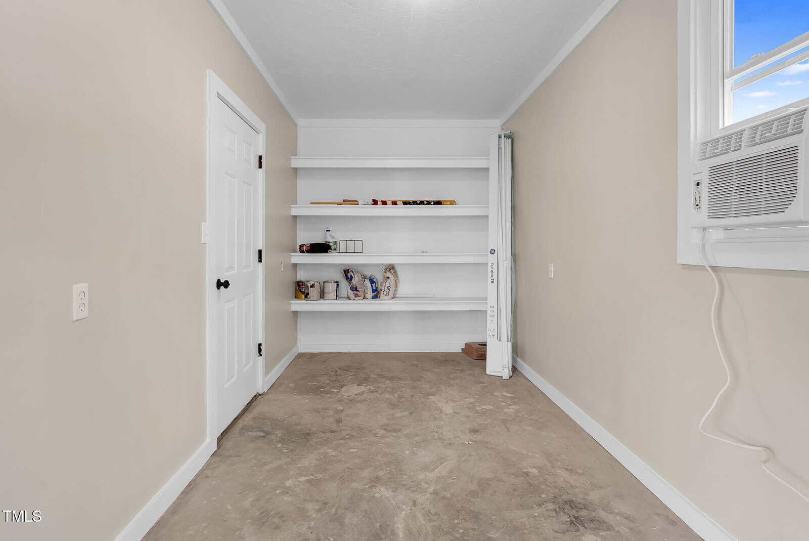A small, empty room with beige walls, an air conditioner in the window, white built-in shelves with a few items, a white door, and an unfinished concrete floor.