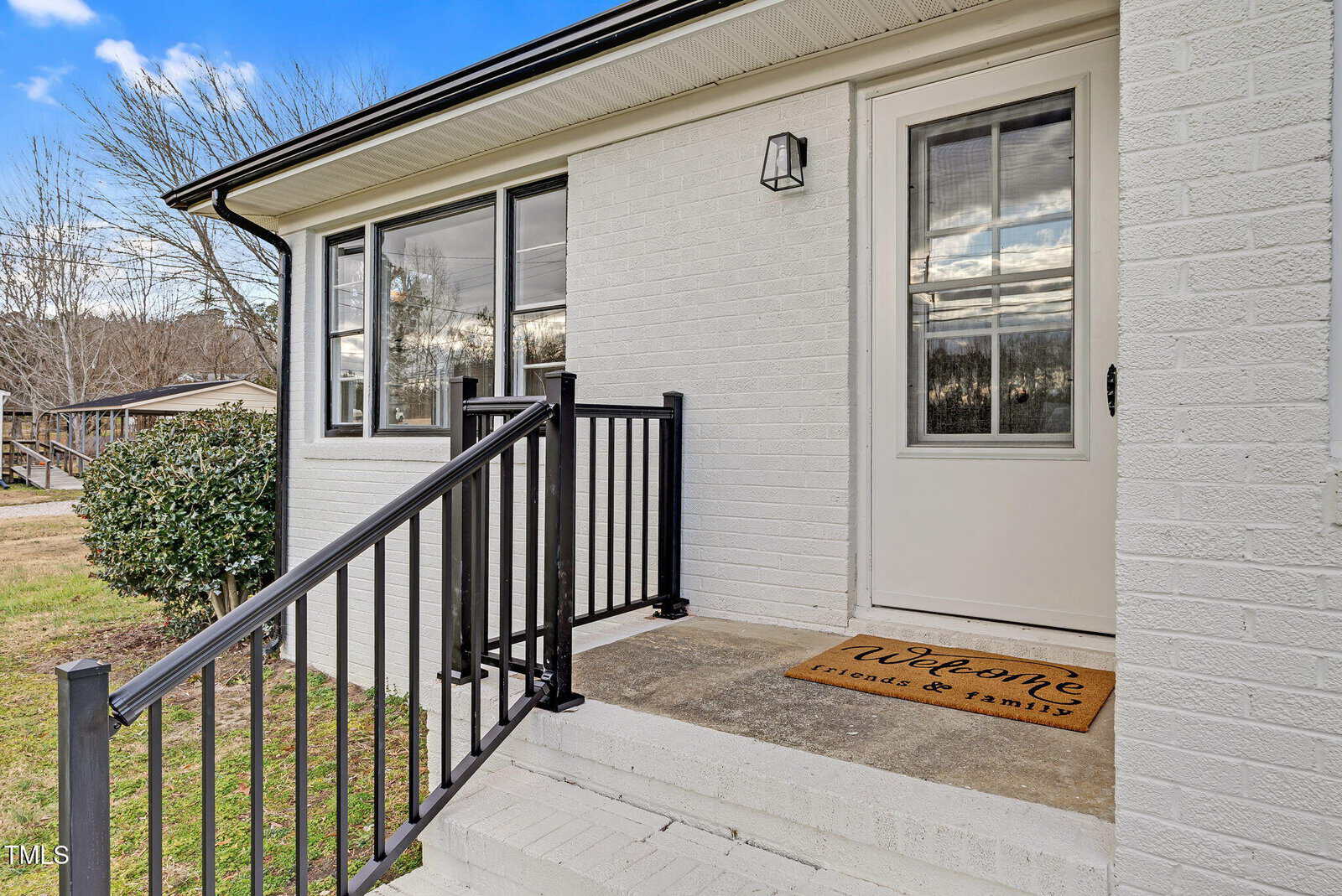 A small porch with black railing leads to a white door on a light brick house. A welcome mat reads "Welcome friends & family." Blue sky and trees are visible in the background.
