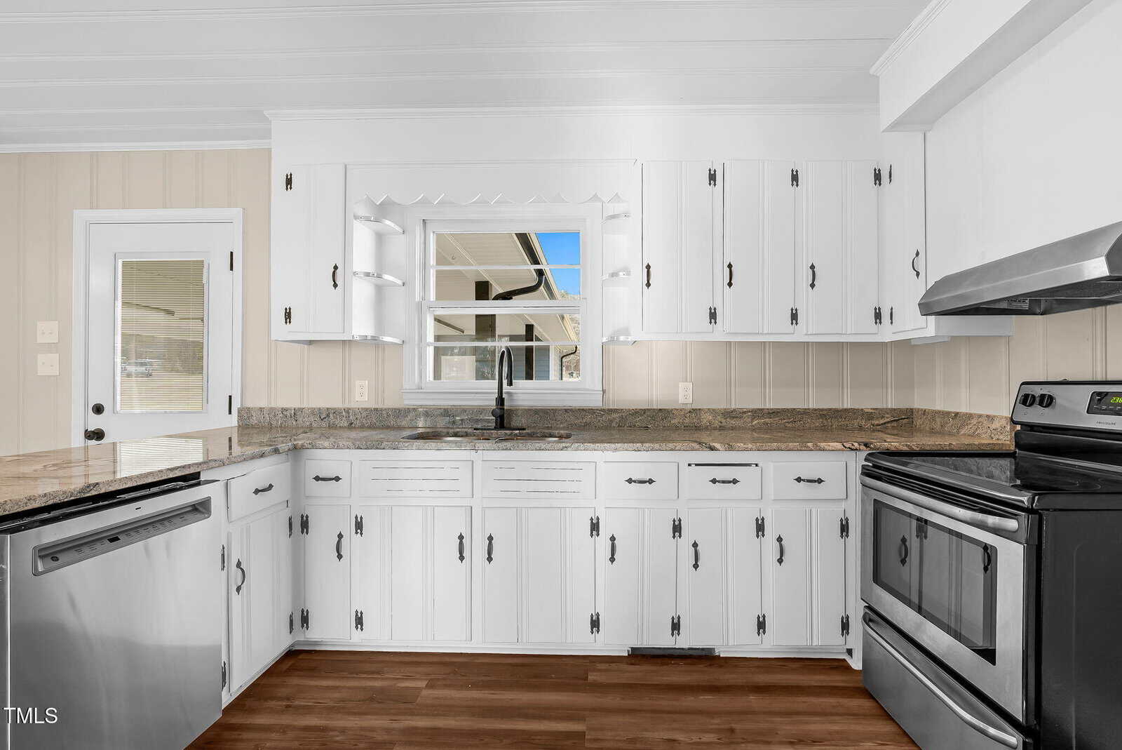 Bright kitchen with white cabinets, granite countertops, stainless steel appliances, and a large window above the sink. Wood flooring and light-colored paneled walls add warmth to the space.