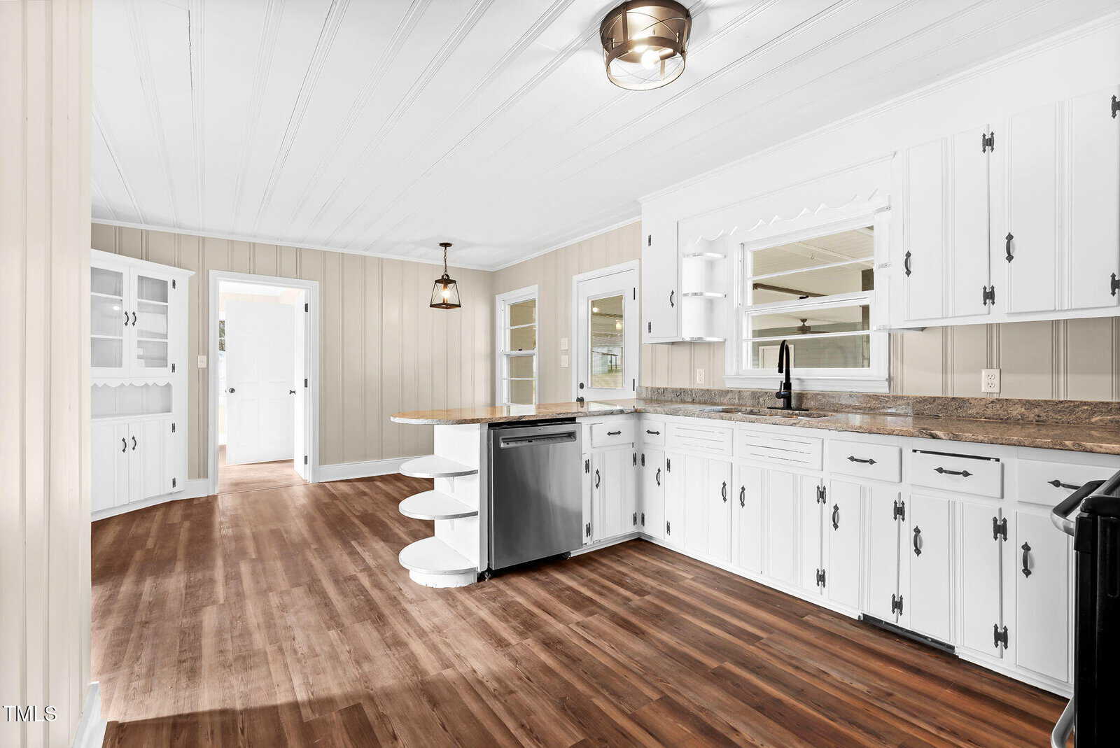 Bright kitchen with white cabinets, wood countertops, a farmhouse sink, stainless steel appliances, and wood flooring. Open shelving and ample windows provide natural light; adjoining dining area visible in the background.