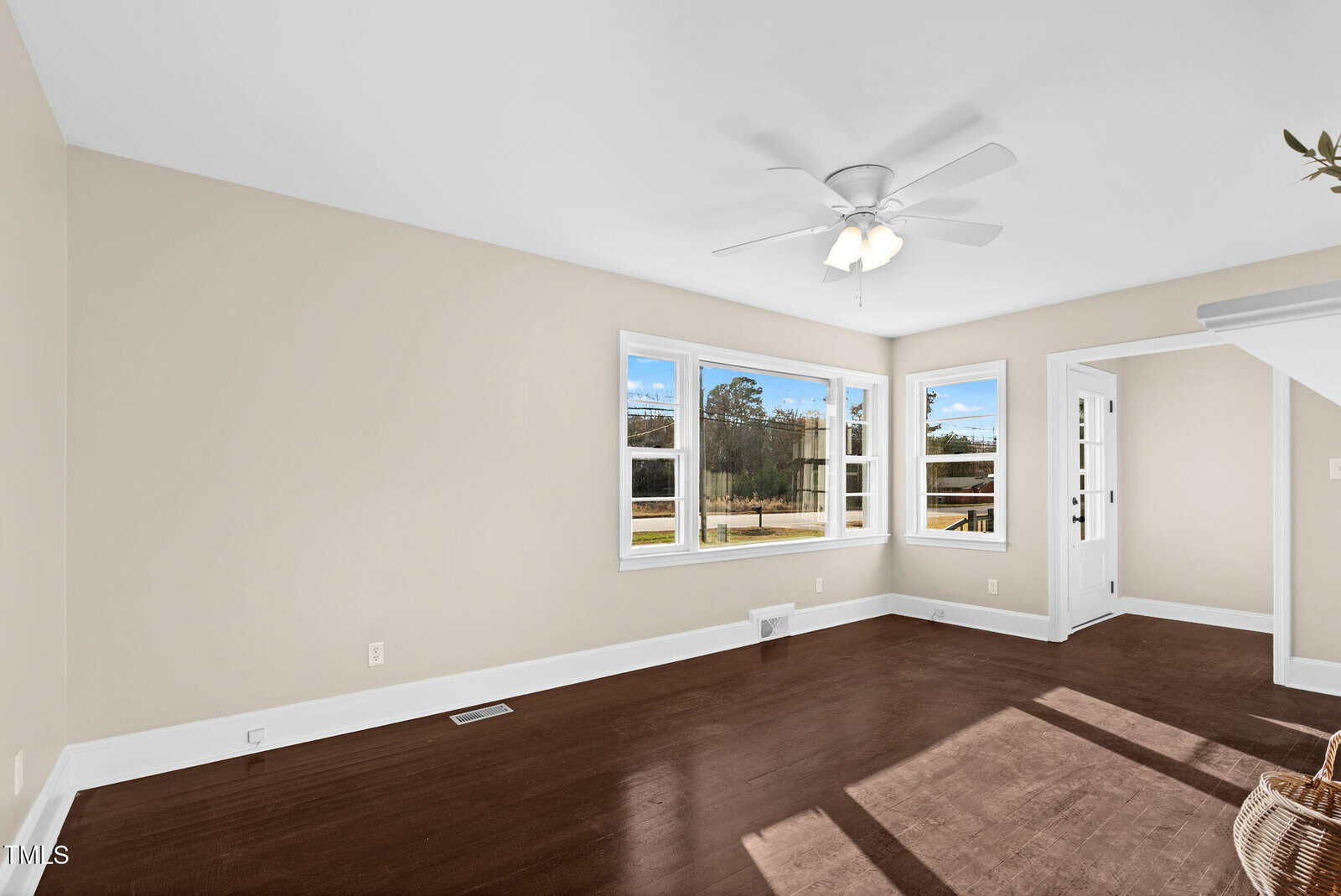A bright, empty room with beige walls, dark wood floors, three large windows, a white ceiling fan, and a small entryway with a white door and more windows.