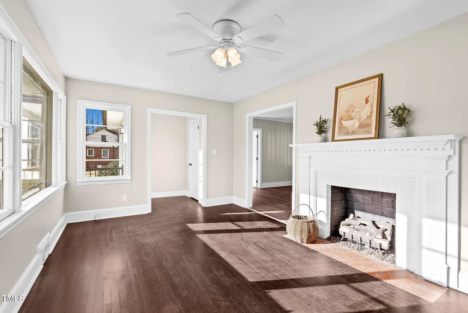 Bright living room with large windows, dark wooden floors, a white ceiling fan, and a white fireplace decorated with artwork and greenery. Sunlight streams in, highlighting the airy, inviting space.