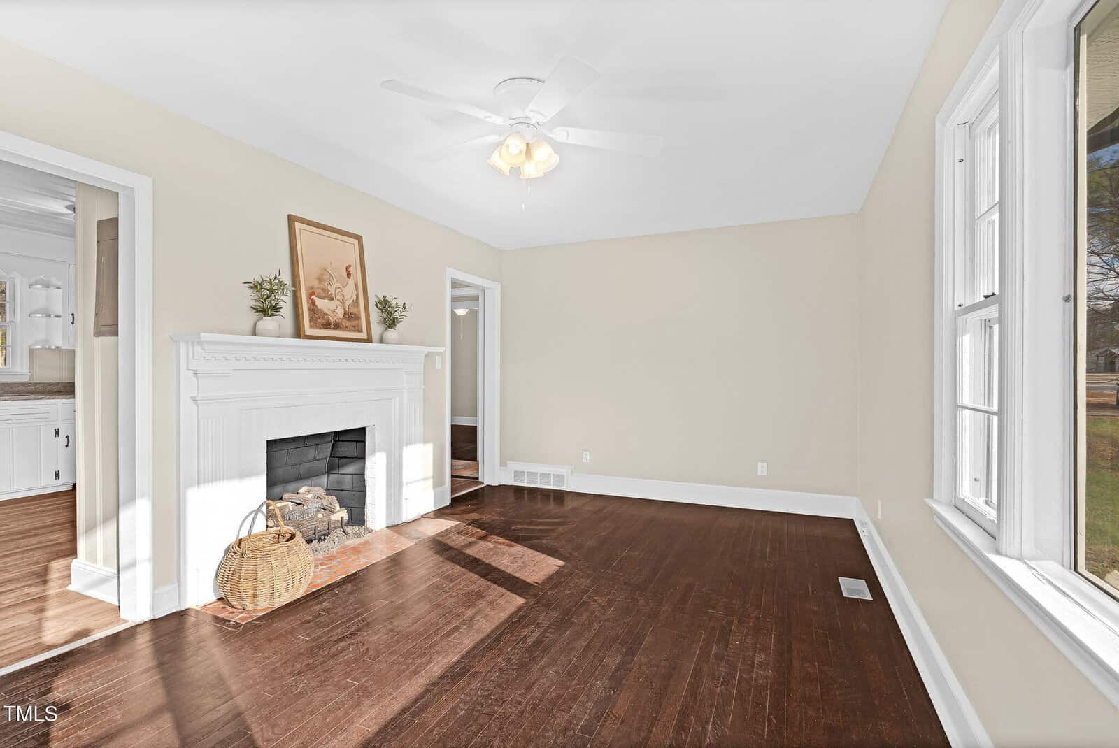Bright living room with dark wood floors, white trim, a white fireplace, artwork above the mantel, a basket with firewood, beige walls, and large windows letting in natural light.
