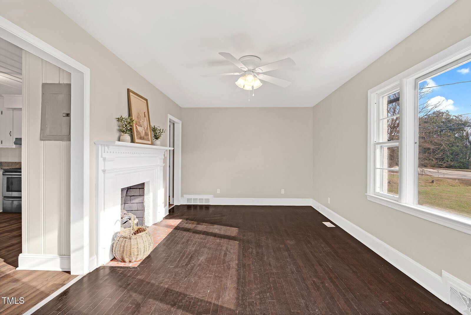 Bright living room with dark wood floors, a white ceiling fan, light gray walls, large windows, and a white fireplace decorated with plants and art. A door opens to the kitchen with white cabinetry.