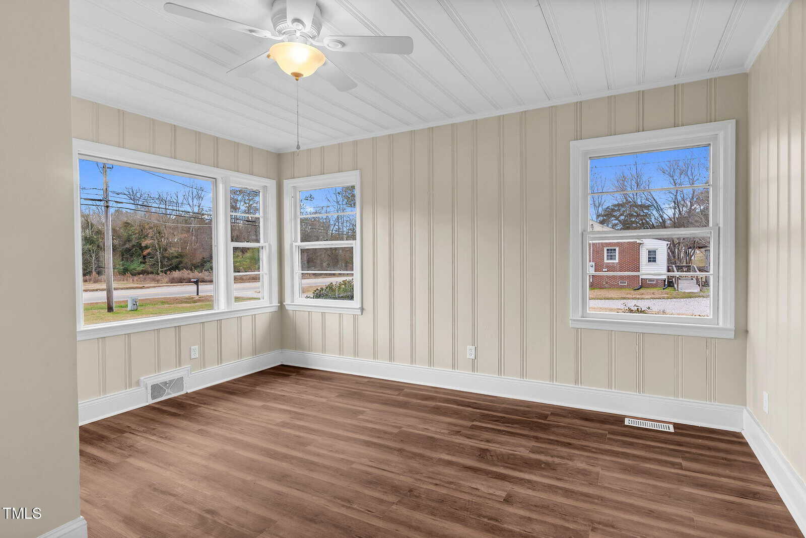 Bright room with beige paneled walls, white ceiling, ceiling fan, and large windows. The wood-style floor contrasts with the neutral tones. The windows offer views of a street, trees, and a neighboring brick house outside.
