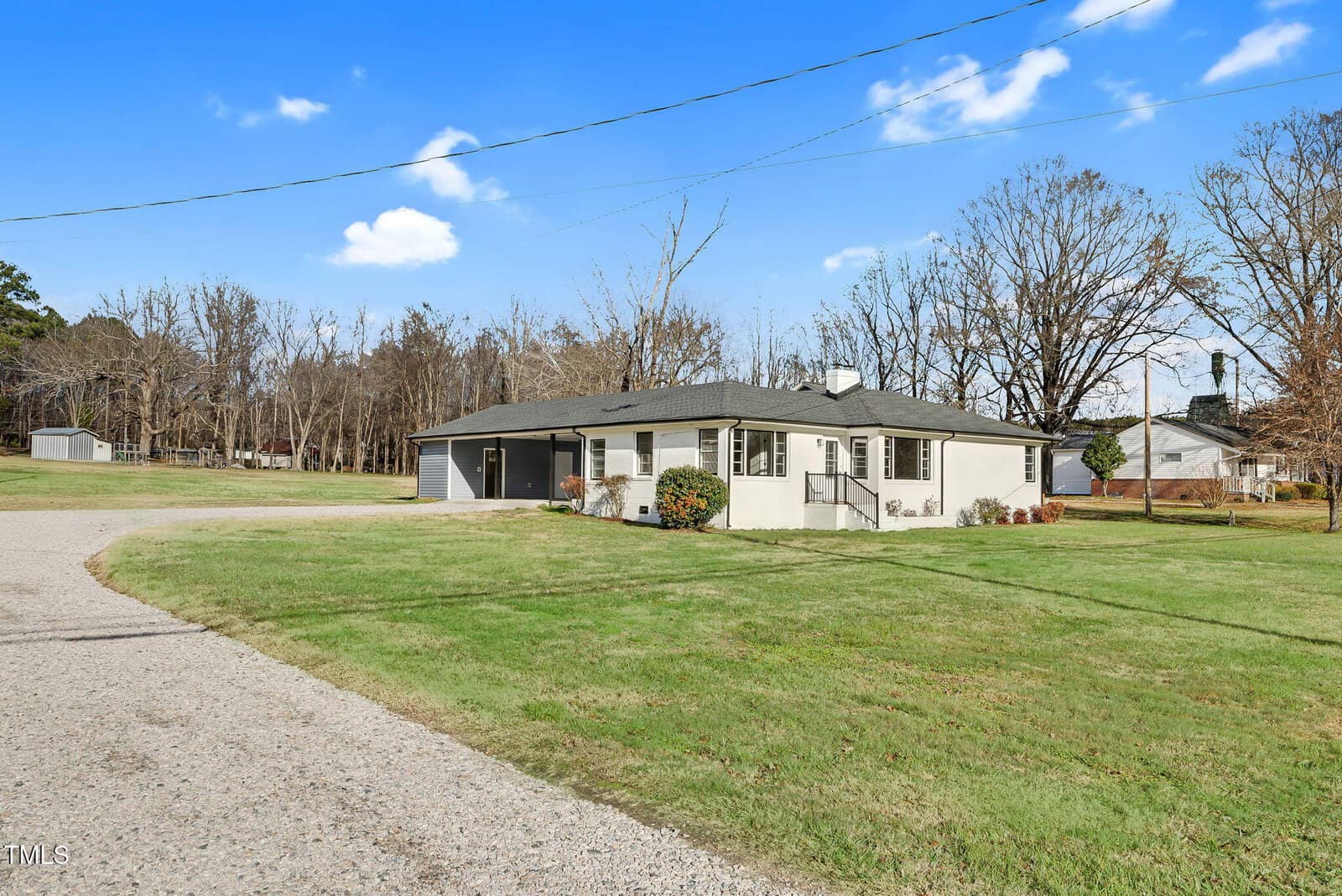 Single-story white house with dark roof at 311 NC 56 Highway E, Louisburg. Features an attached carport, large lawn, trees with bare branches in back, blue sky with scattered clouds, and a gravel driveway curving toward the house.