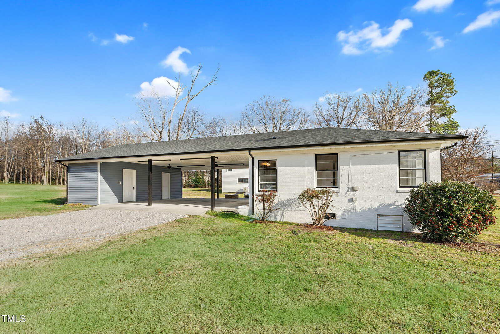 Single-story white house with a dark roof, attached carport, gravel driveway, and neatly trimmed grass lawn under a blue sky with a few clouds. Trees are visible in the background.