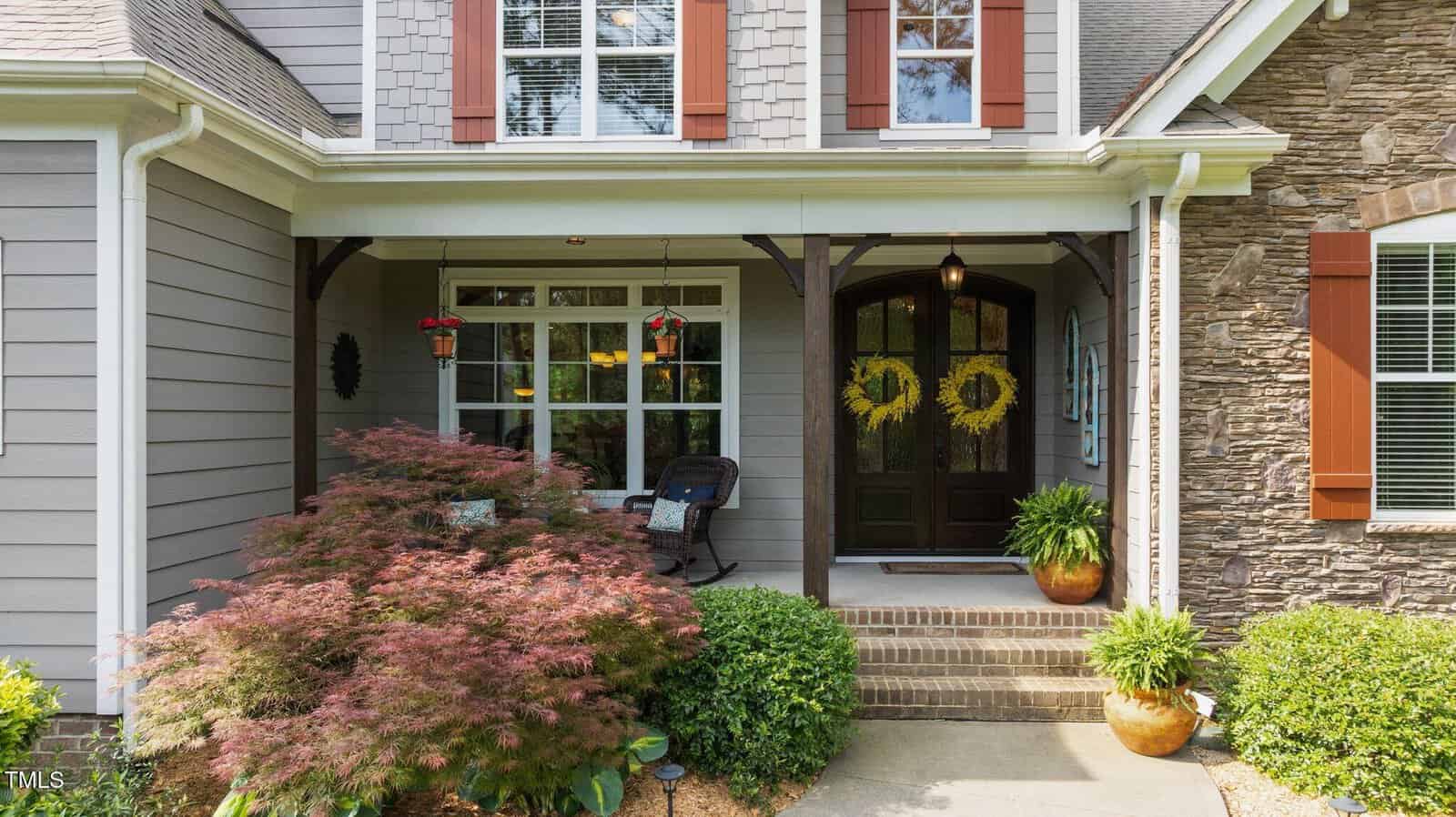 A cozy house front porch with double wooden doors decorated with yellow wreaths, a wicker chair, potted plants, hanging flowers, and a Japanese maple tree near the entrance. The home exterior has gray siding and stone accents.