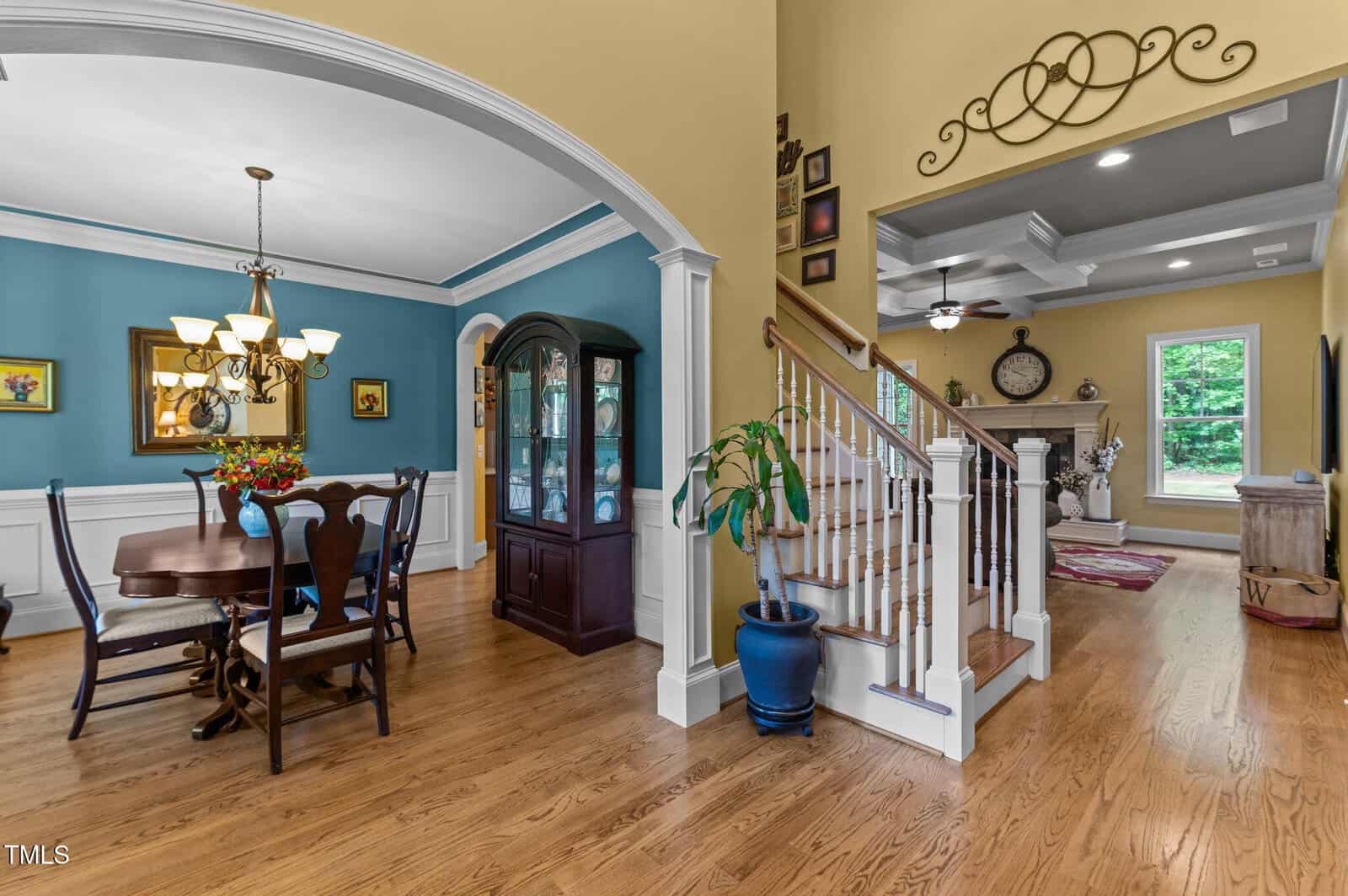 Open dining area with a wooden table and chairs, blue accent wall, chandelier, china cabinet, and adjacent staircase with wood and white railings. Living area with fireplace is visible in the background.