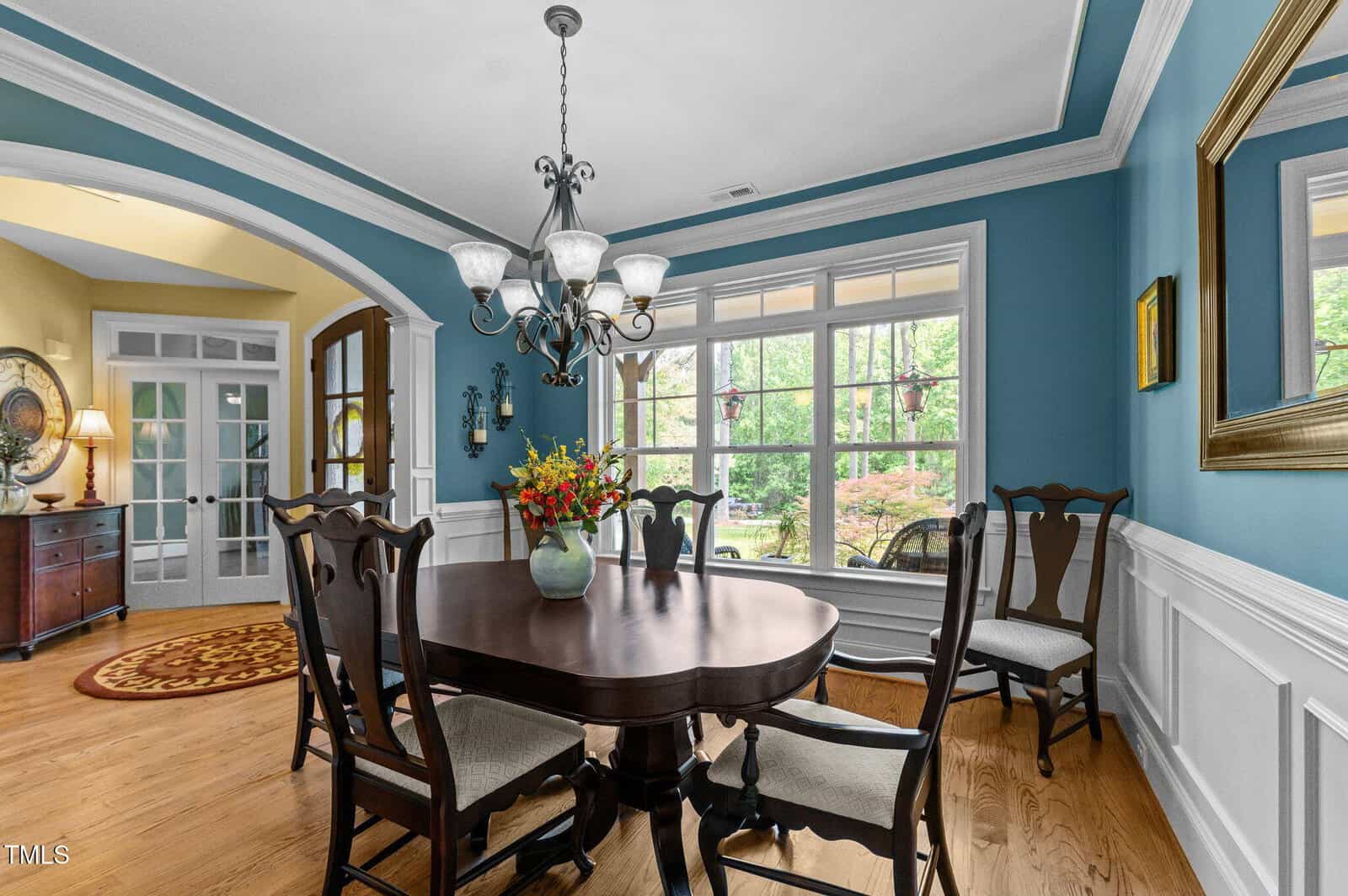 A dining room with blue walls, white wainscoting, a dark wood table and chairs, a chandelier, a large window with a view of greenery, and a vase of flowers on the table.