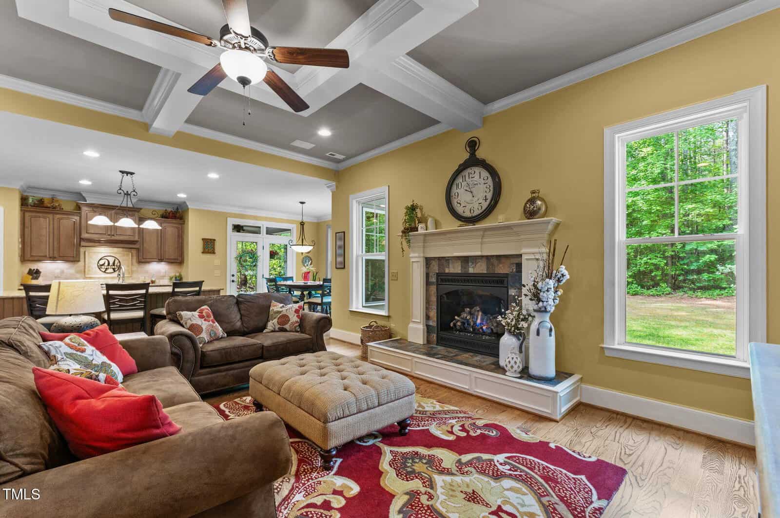 A cozy living room with brown sofas, colorful pillows, a tufted ottoman, a fireplace with a clock and vases on the mantel, wood floors, yellow walls, large windows, and a view into the kitchen and dining area.