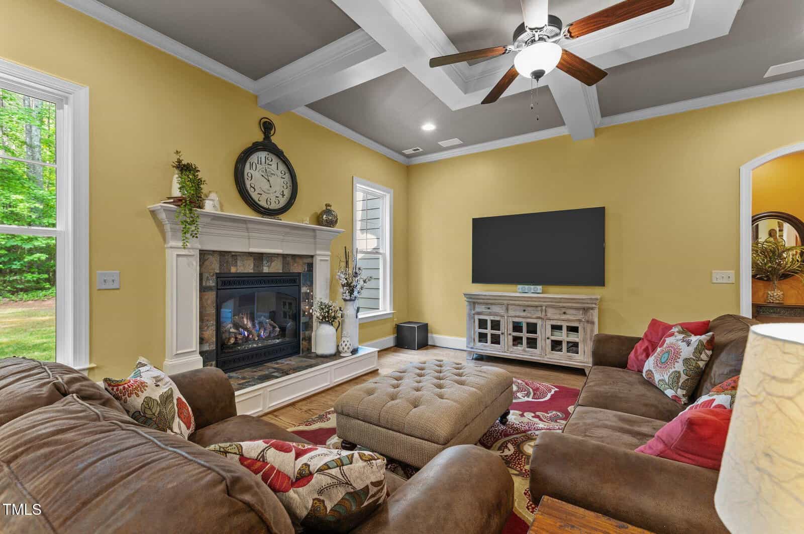 Living room with yellow walls, sectional sofa, ottoman, fireplace with clock, TV on console, ceiling fan, and large windows showing greenery outside. Patterned rug and decorative plants are also visible.