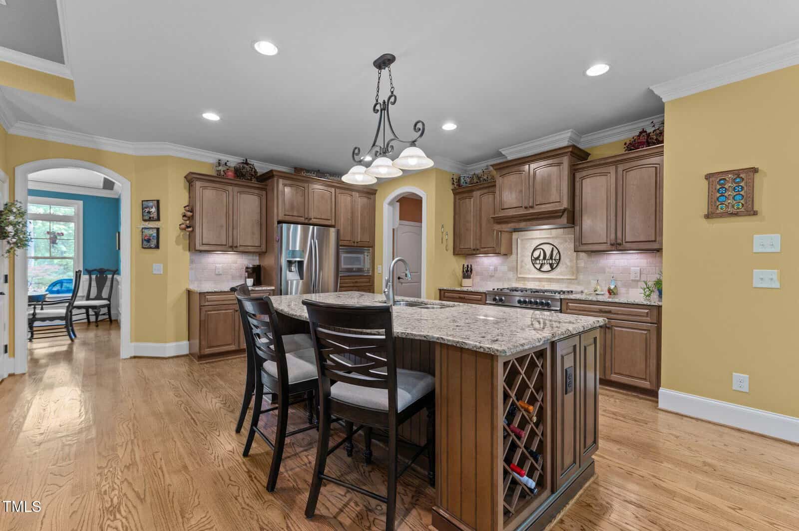 Spacious kitchen with wood cabinets, granite island with built-in wine rack, two chairs, stainless steel appliances, recessed lighting, and yellow walls. An arched doorway leads to another room with large windows.