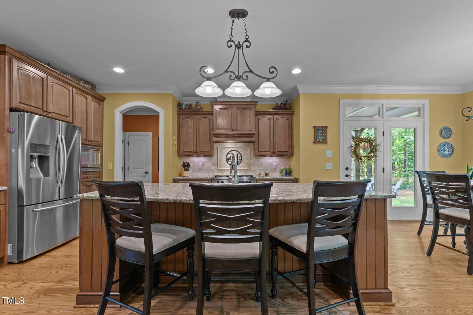 Modern kitchen with wooden cabinets, stainless steel appliances, an island with granite countertop, and four dark wooden chairs. Pendant light fixture hangs above the island. Glass doors and windows show a green outdoor area.