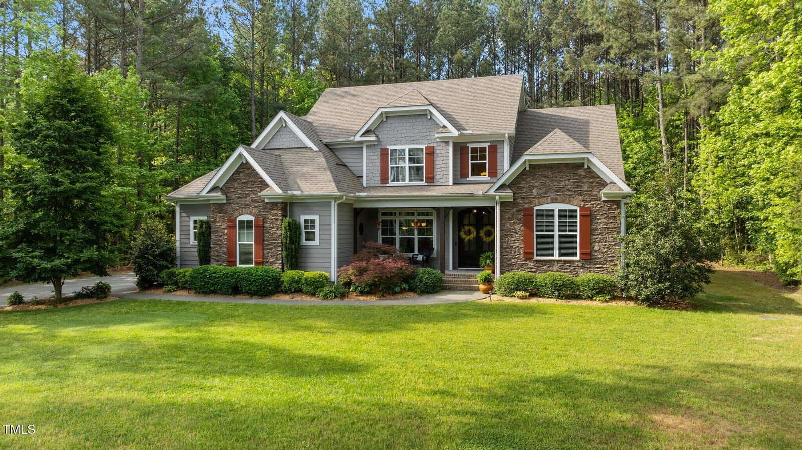 A two-story house with gray siding and brick accents, featuring white trim, multiple gables, red shutters, a covered front porch, and a well-manicured lawn, surrounded by trees and greenery.