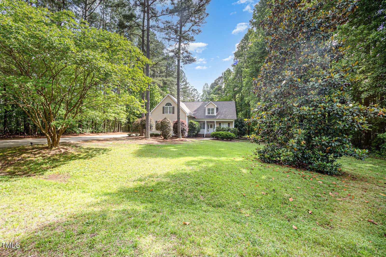 A white house with a gabled roof sits surrounded by tall trees and a large, grassy front yard under a bright blue sky with scattered clouds. Bushes and mature trees frame the driveway and the house.