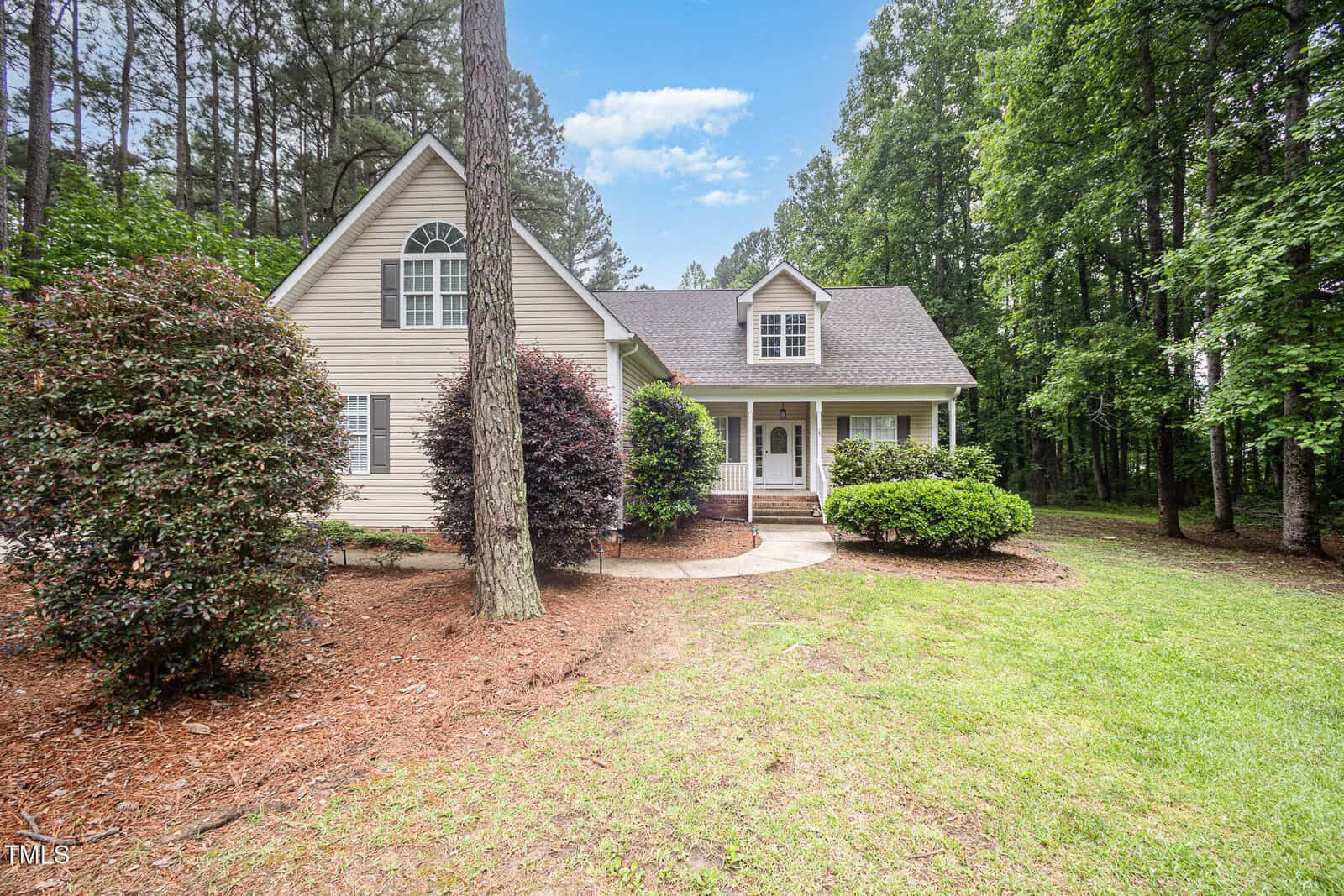A beige house with a covered front porch, white trim, and steep roof sits among tall trees and bushes, with a curved walkway leading to the entrance and grass lawn in front.