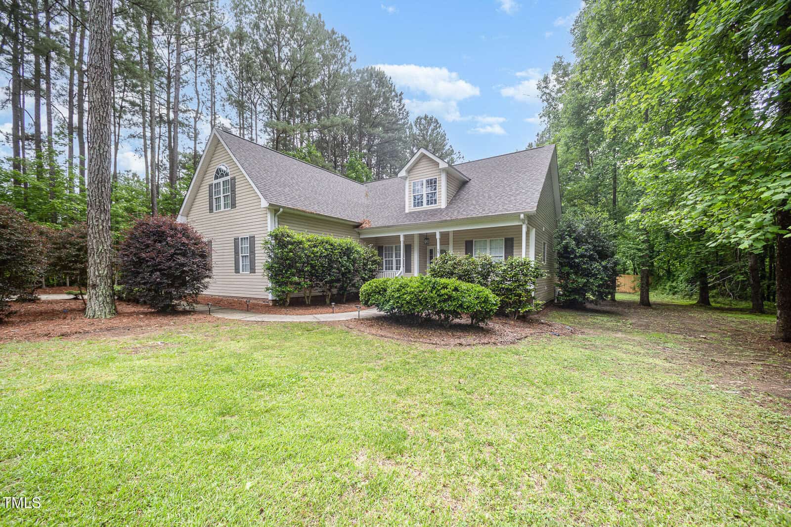 A beige two-story house with a gabled roof sits surrounded by tall trees and lush greenery. Well-maintained bushes line the front, and a large grassy lawn stretches out in front of the home.