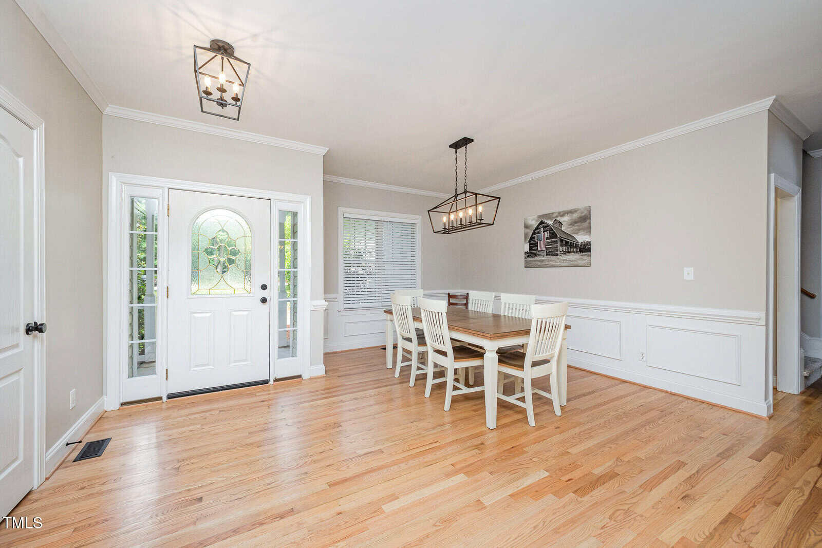 A bright dining room with light wood floors, white walls, a wooden dining table with six white chairs, a modern chandelier, a large window, and a white front door with glass panels.