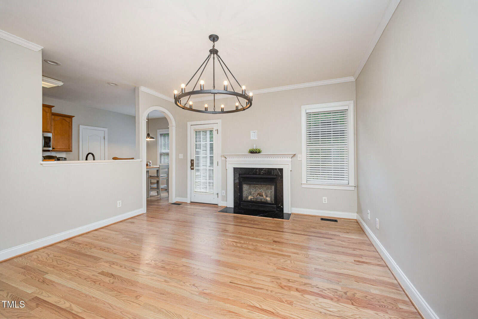 Bright, empty living room with light wood floors, a black chandelier, white walls, a black fireplace, two windows, and a door leading outside. The kitchen with wood cabinets is partially visible through an open archway.