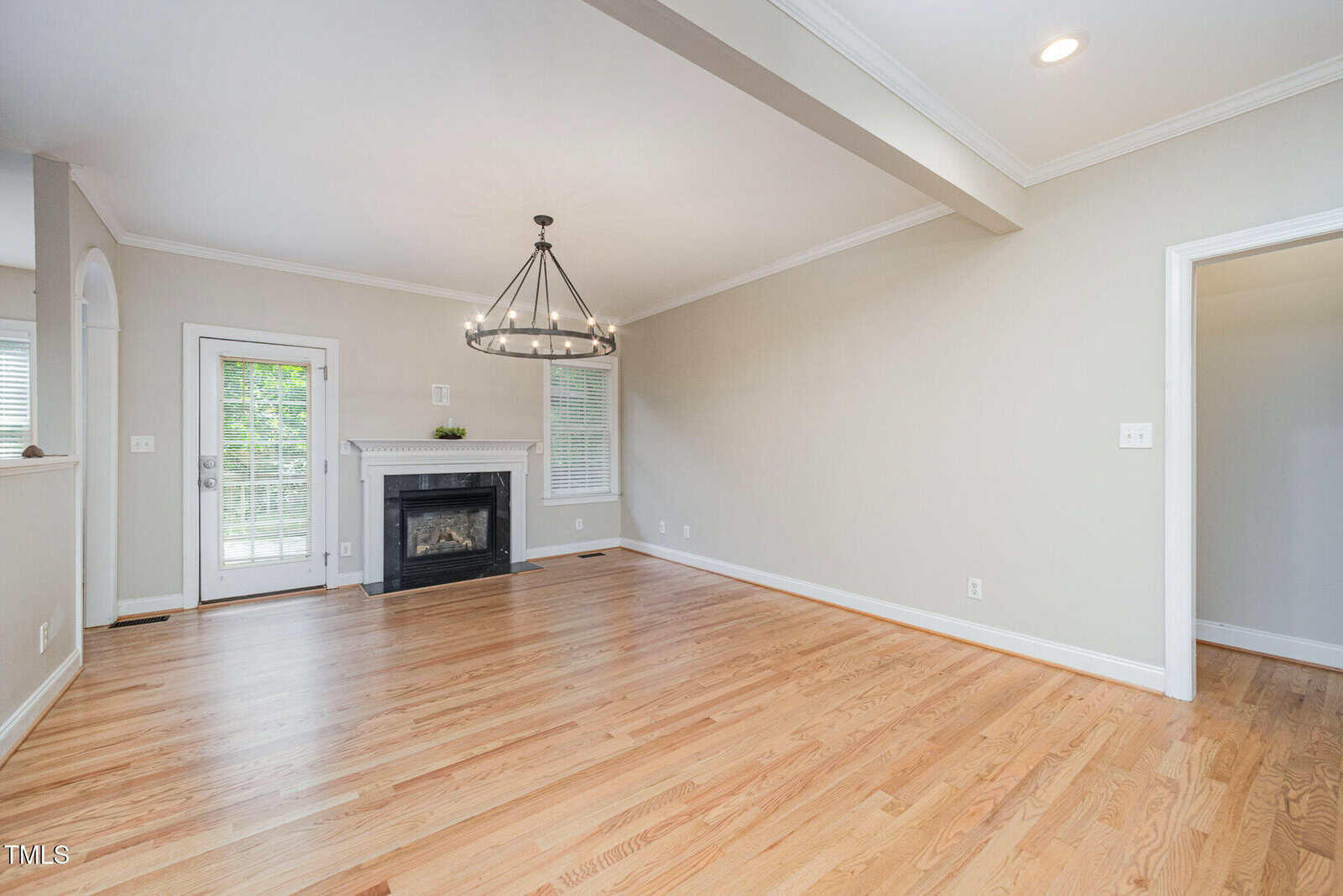 A bright empty living room with hardwood floors, a fireplace, light gray walls, a large chandelier, a window with blinds, and a door leading outside. White trim and ceiling lights add contrast to the neutral decor.